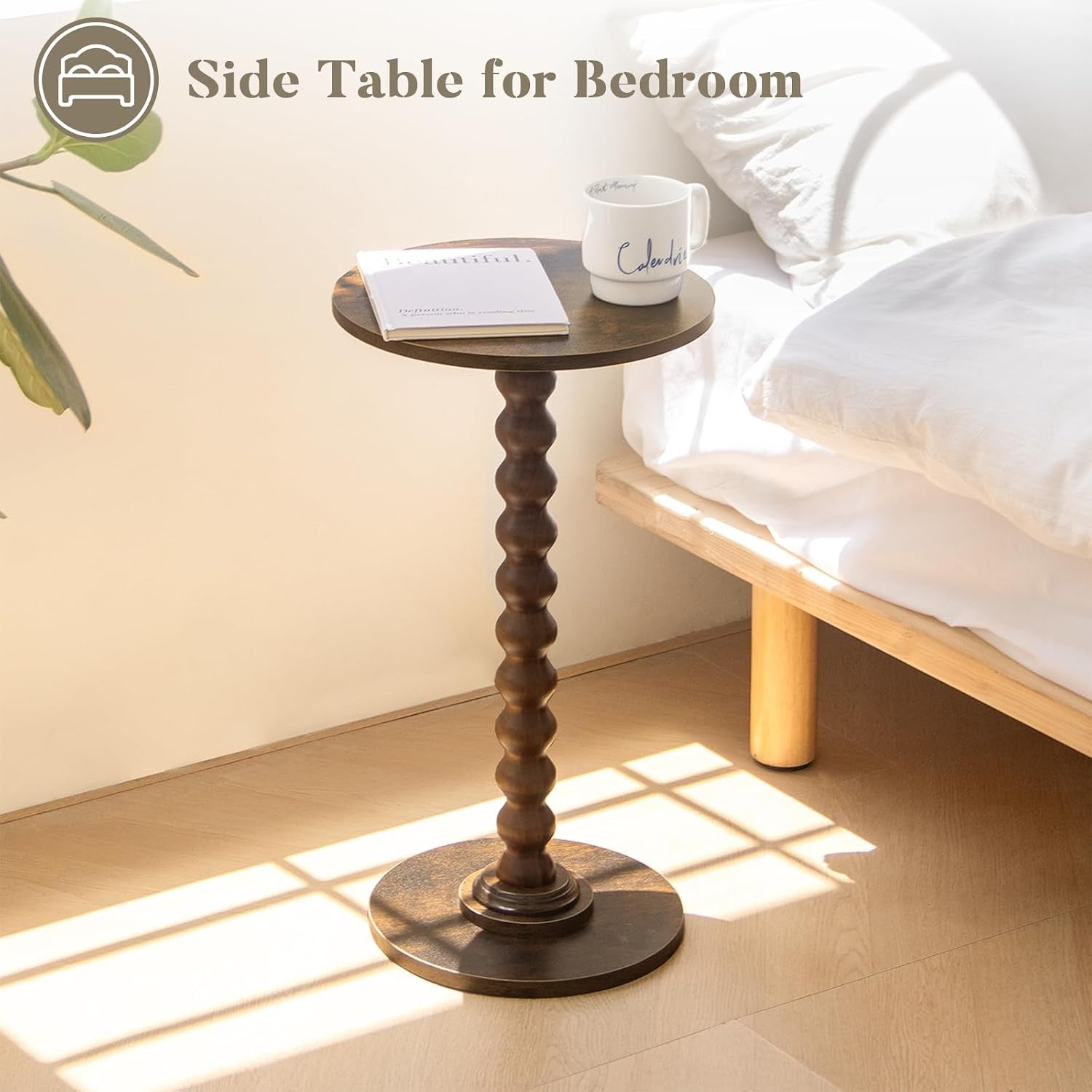 Side table placed beside a low bed in sunlight, holding a mug and a small book on the walnut round top.