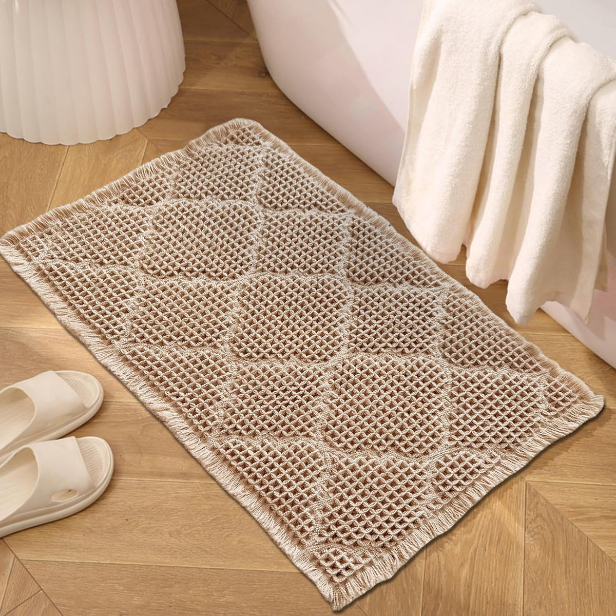 Tan waffle bath mat placed on wood floor beside a bathtub with towel and white slide sandals visible