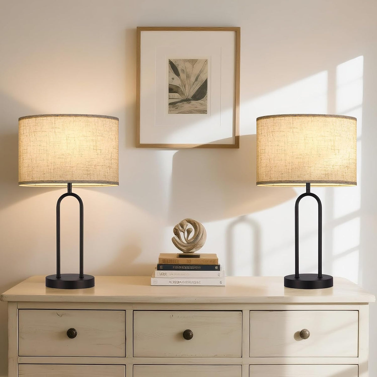 Pair of lit beige oval shades on matching black-arched table lamps atop a cream dresser with framed art between them