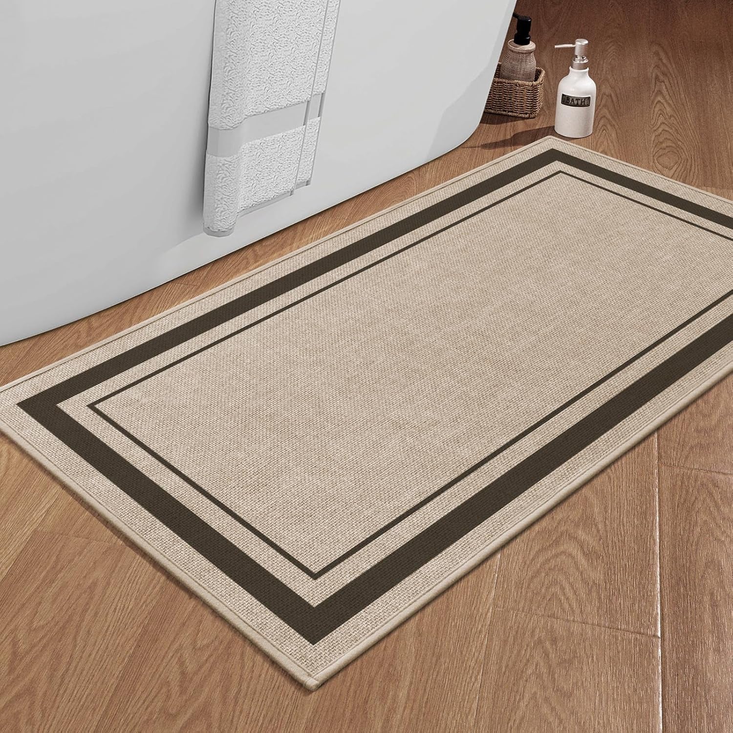 Close angled view of a beige bath mat with dark border placed on wood floor by a bathtub exterior.