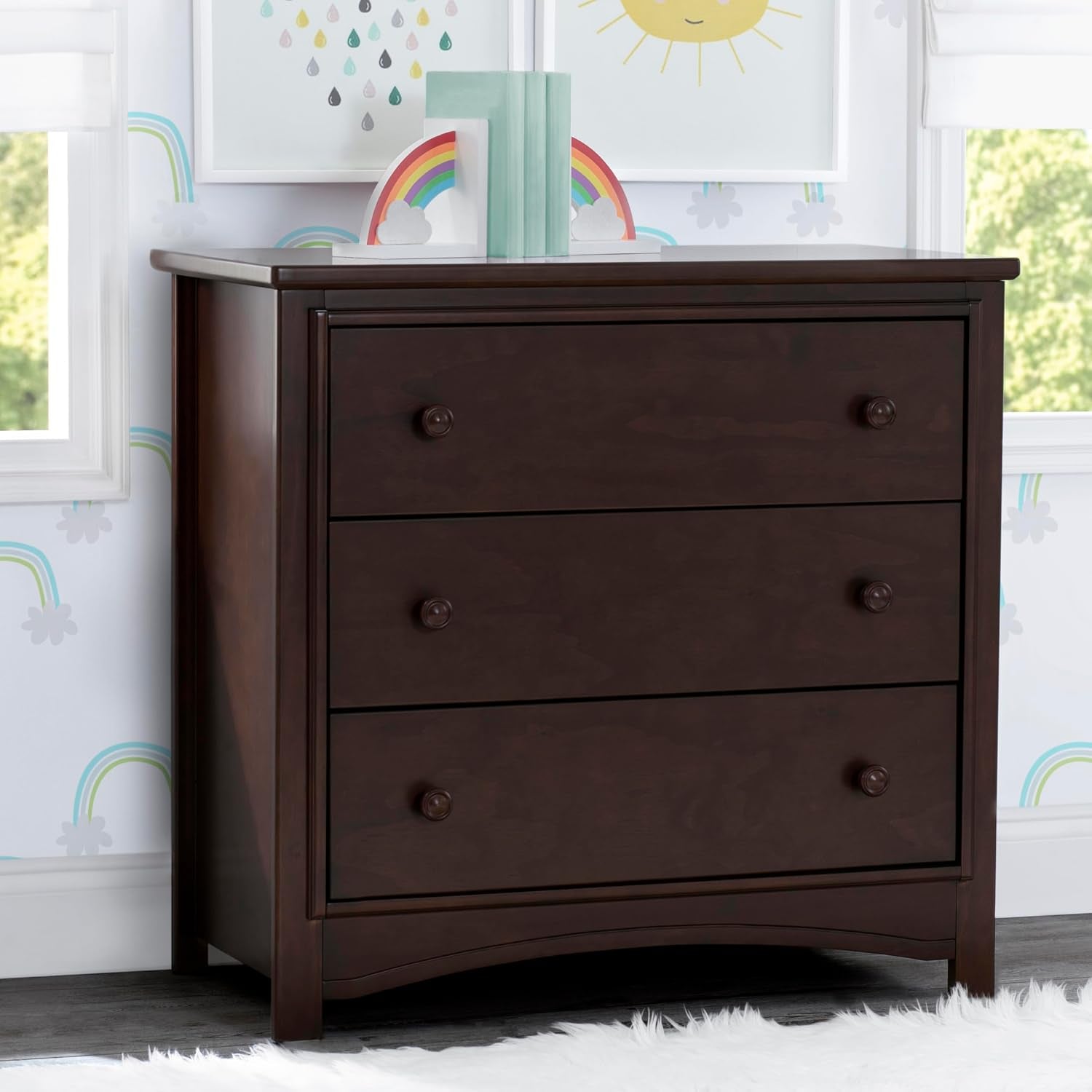 Front view of dresser in a nursery with books and rainbow decor on top and a white shag rug beneath.
