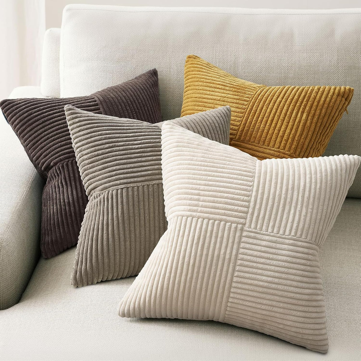 Four ribbed velvet throw pillows in mustard, dark brown, taupe and cream arranged on a light fabric sofa.