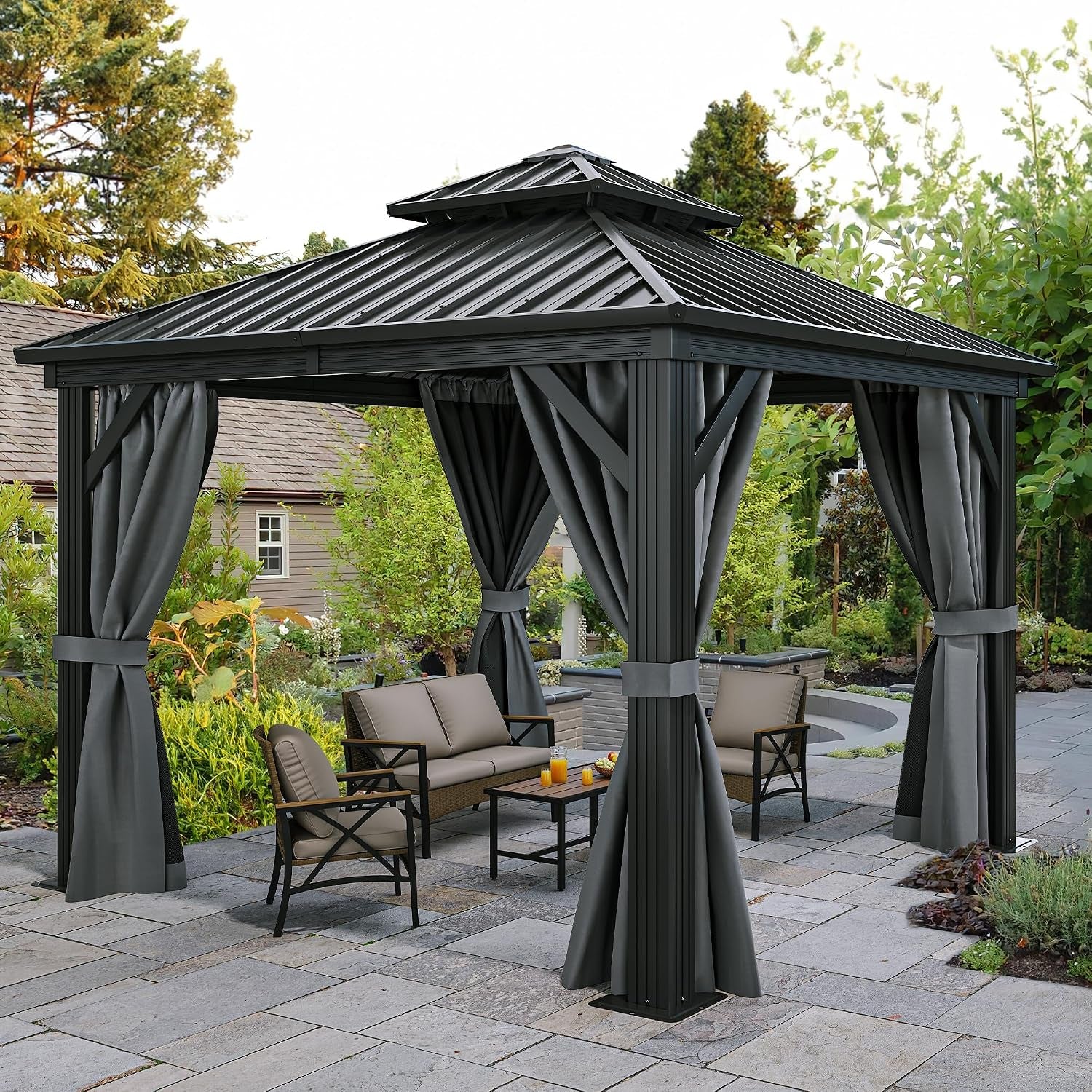 Hardtop gazebo with tied-back gray curtains on a stone patio surrounded by garden greenery