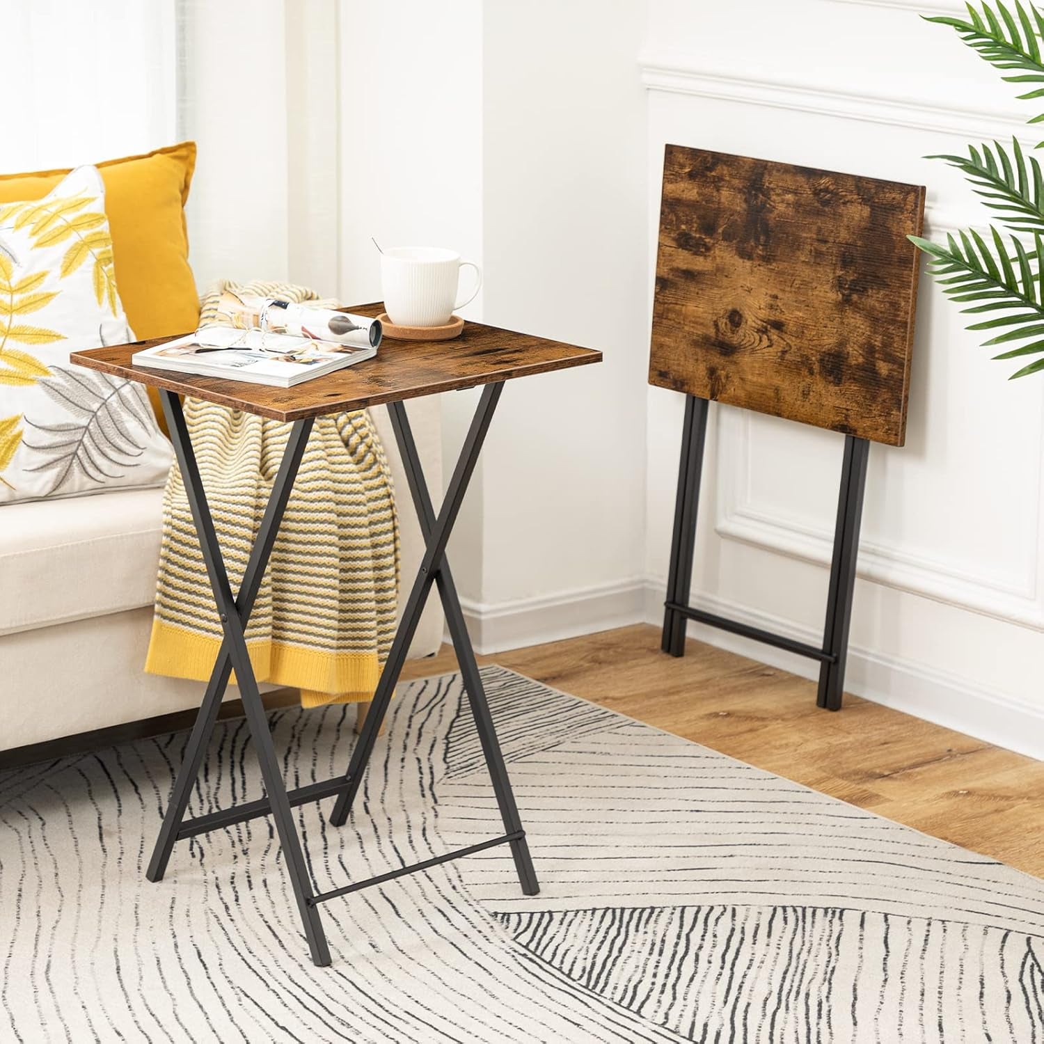 Single folding tray table set up beside a sofa with a mug and magazine, and a second table folded and leaned against the