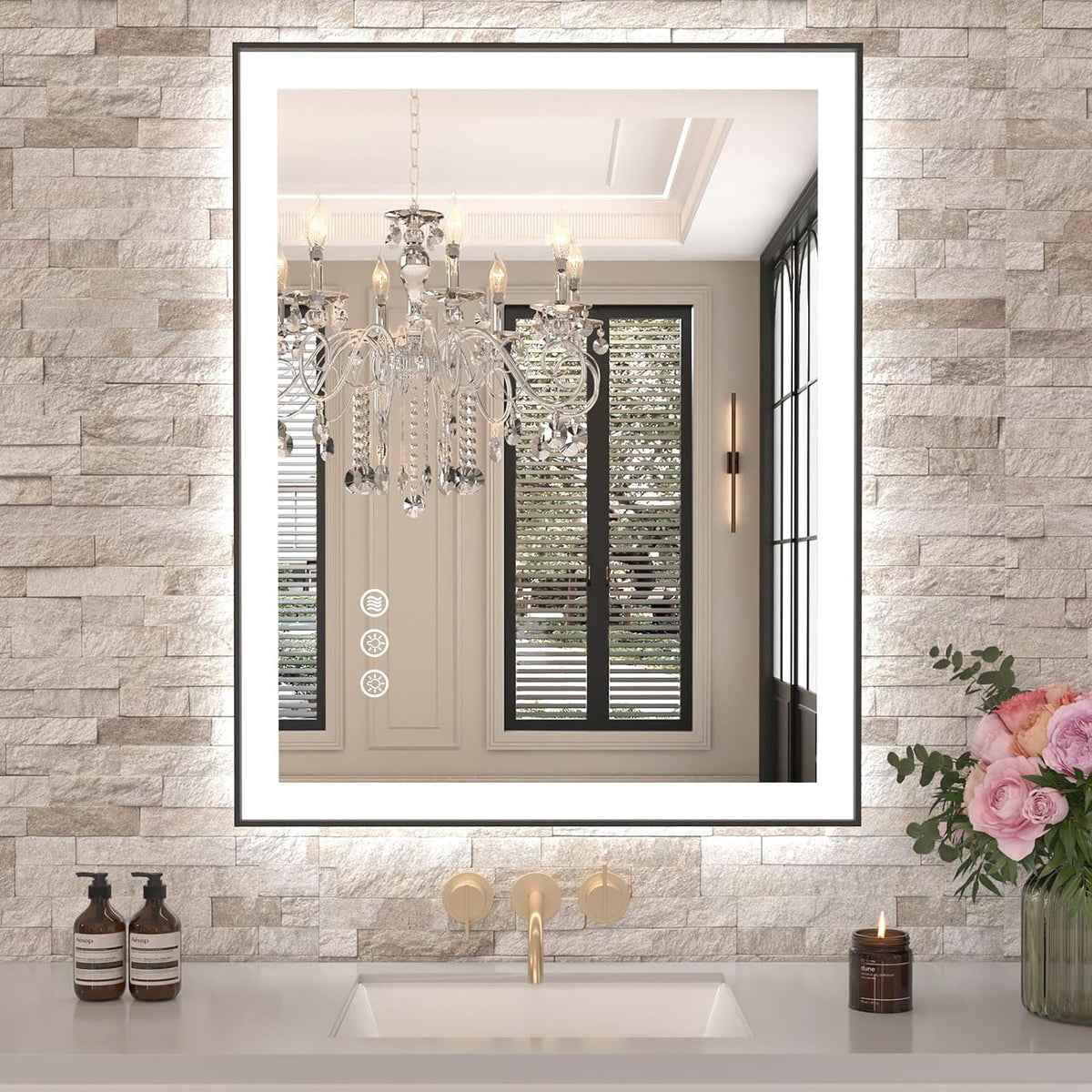Framed LED mirror on textured stone wall reflecting a chandelier, gold faucet and floral arrangement at right