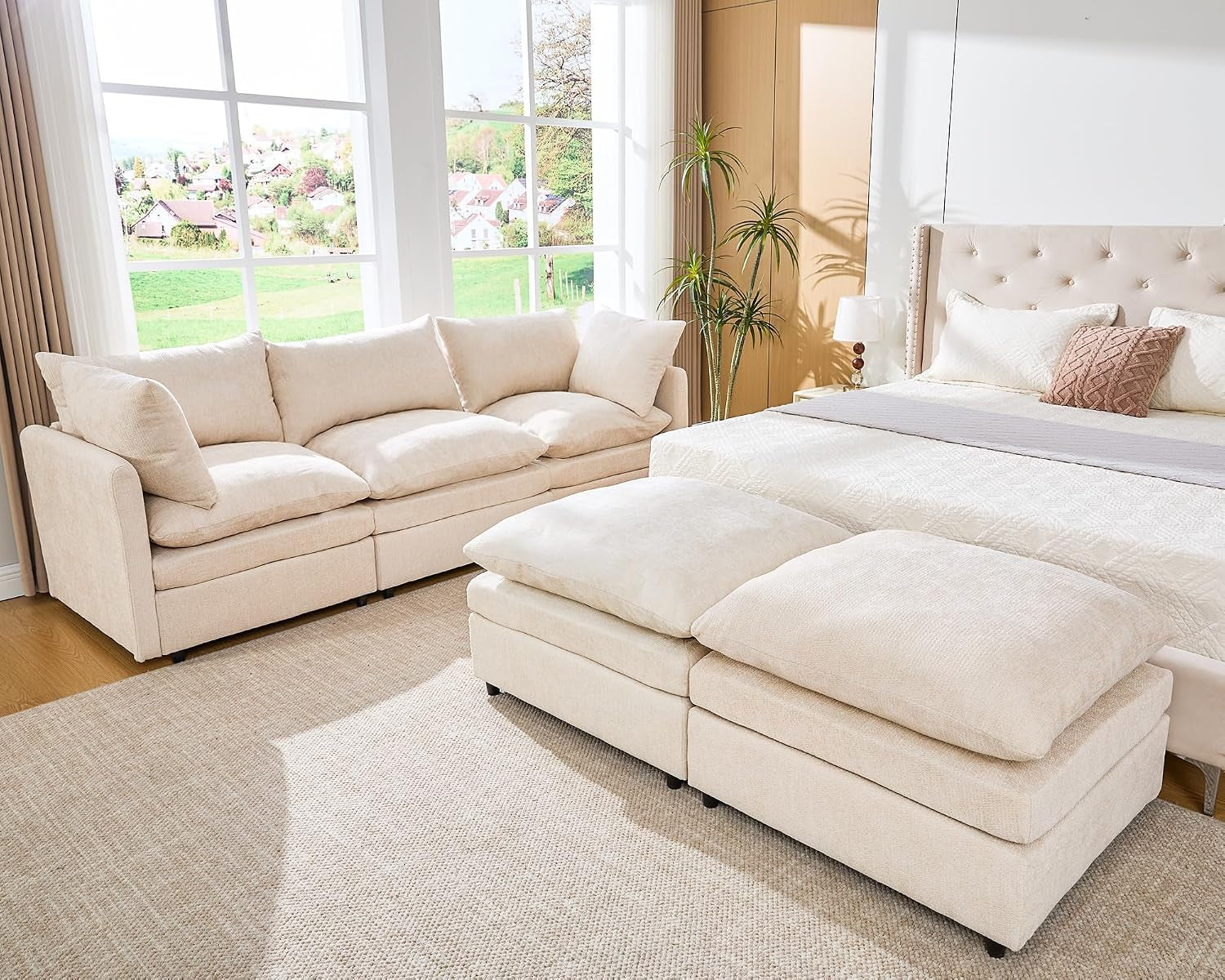 Cream three-seat sofa and two ottomans arranged beside a bed in a bright room.