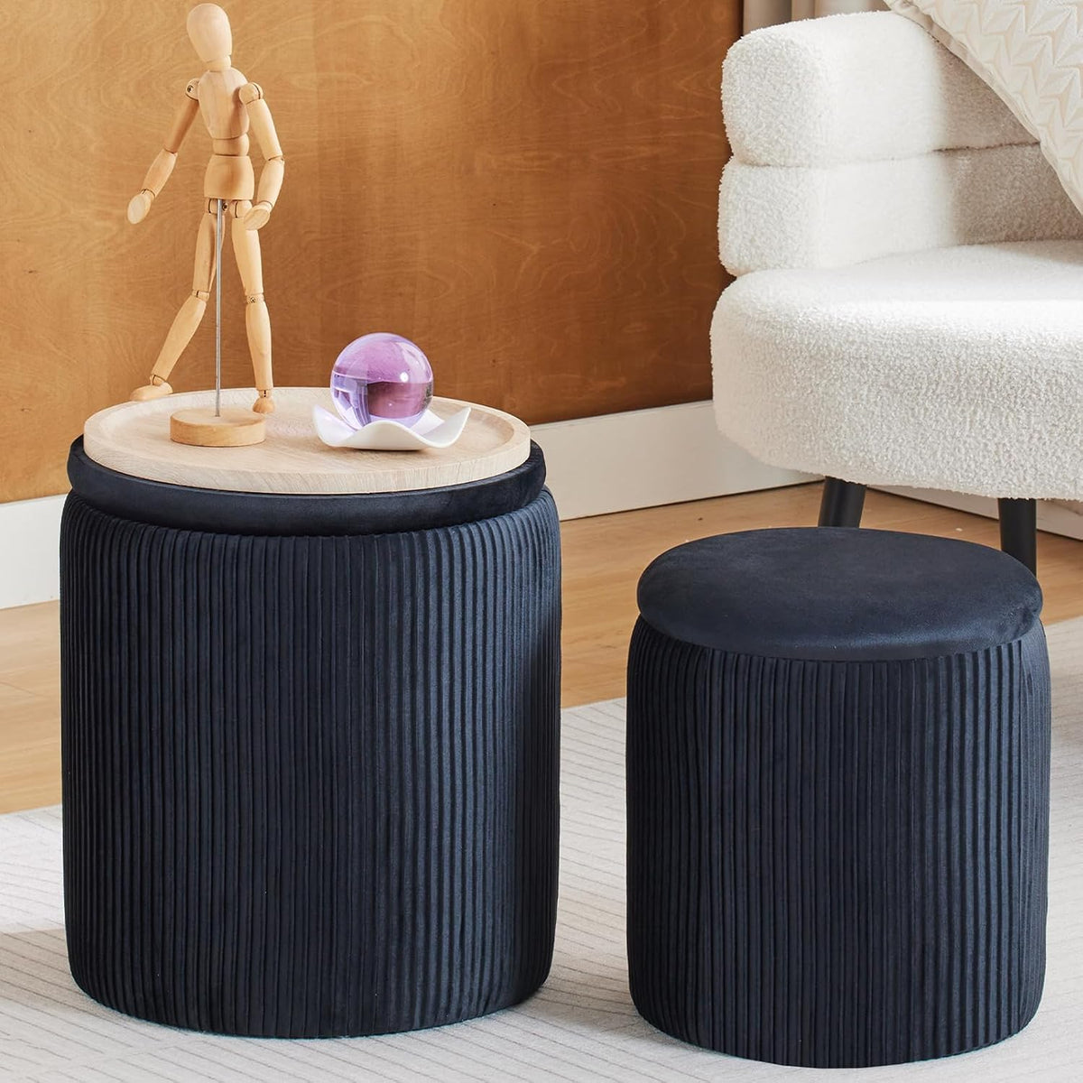 Close angled view of black pleated ottomans, larger topped with a wooden tray holding a decorative orb beside a plush white