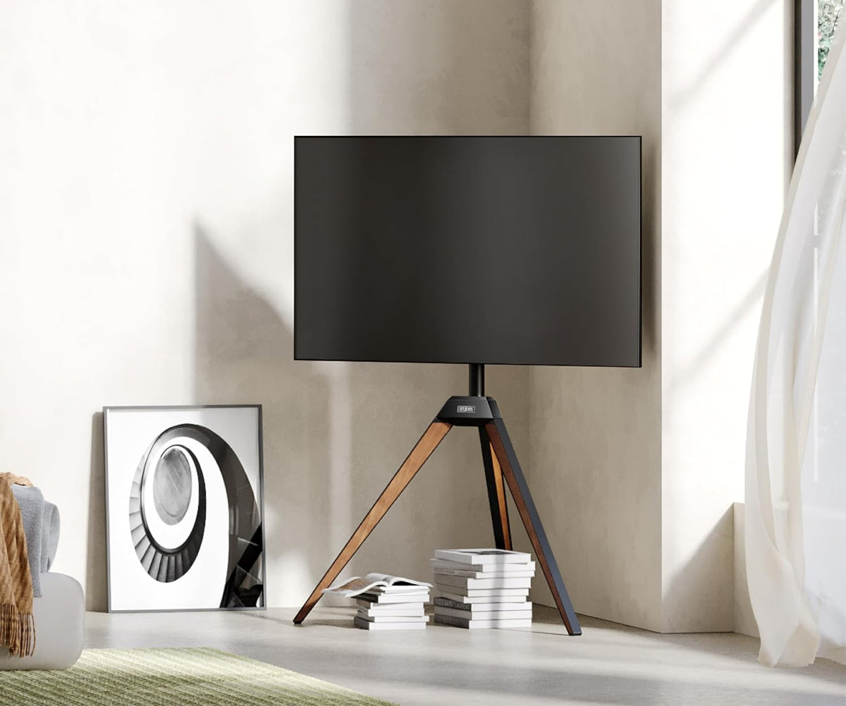 Tripod-mounted TV in a neutral living space beside stacked books and framed artwork leaning on the floor.