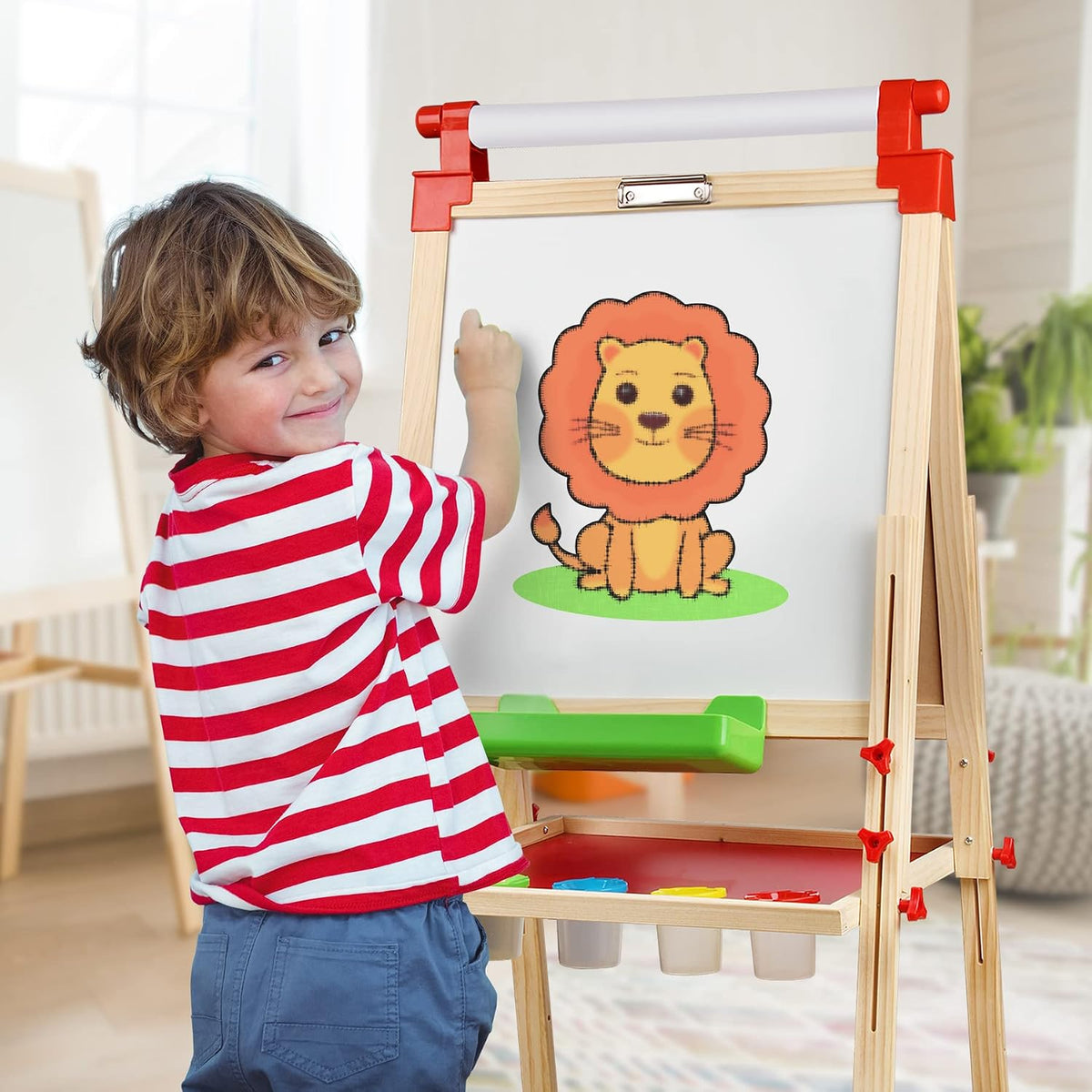 Child in red-striped shirt drawing a lion on a wooden easel with green tray and paint pots underneath.
