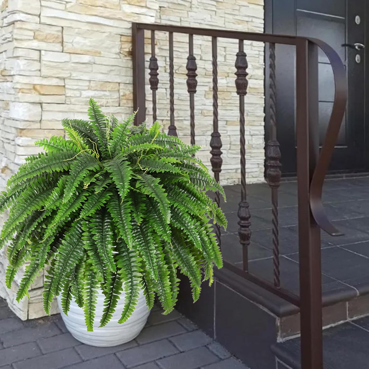 Large artificial Boston fern in a white ceramic pot sitting on a front porch step beside a brown metal railing.