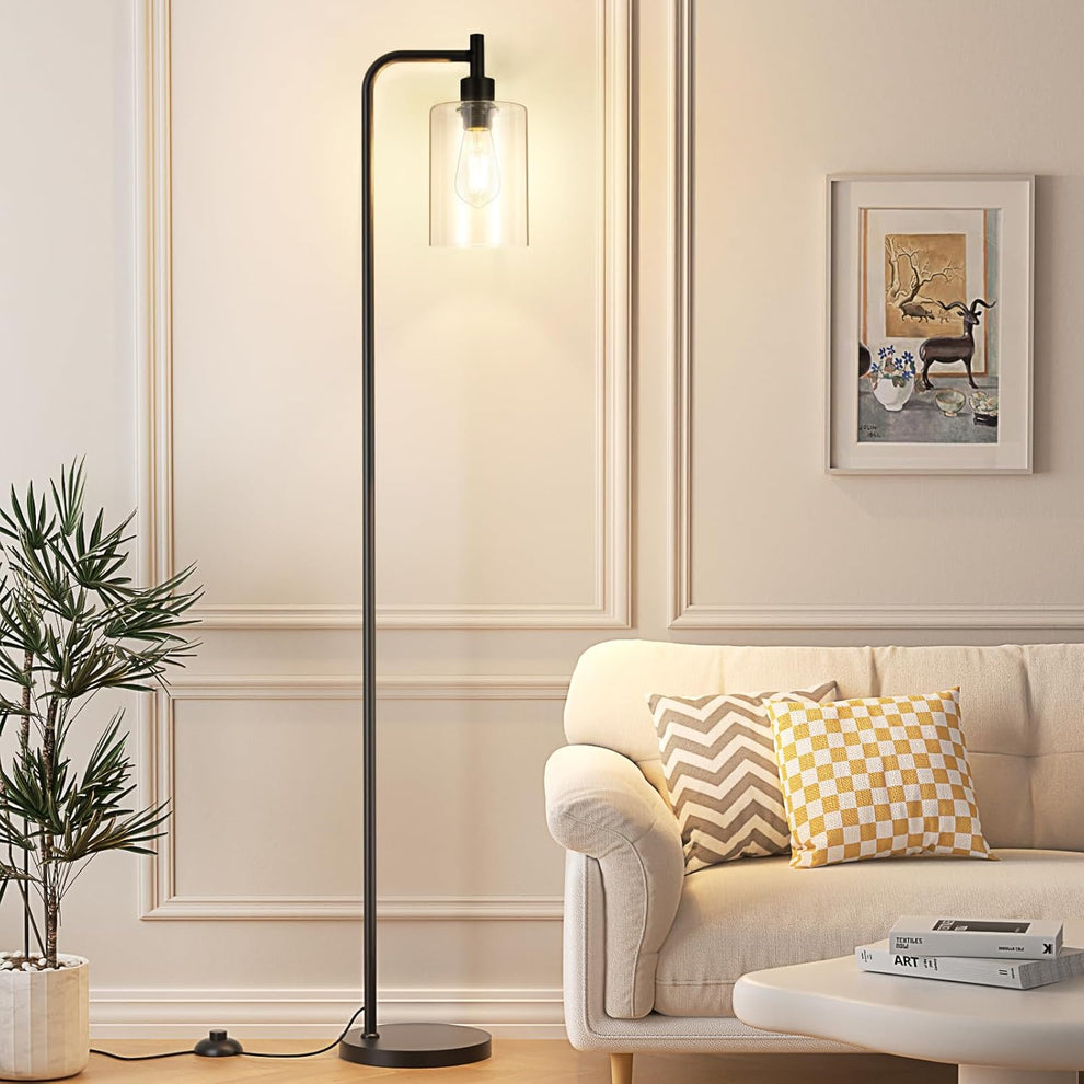 Slim black floor lamp with clear cylindrical glass shade next to a beige sofa, angled front view