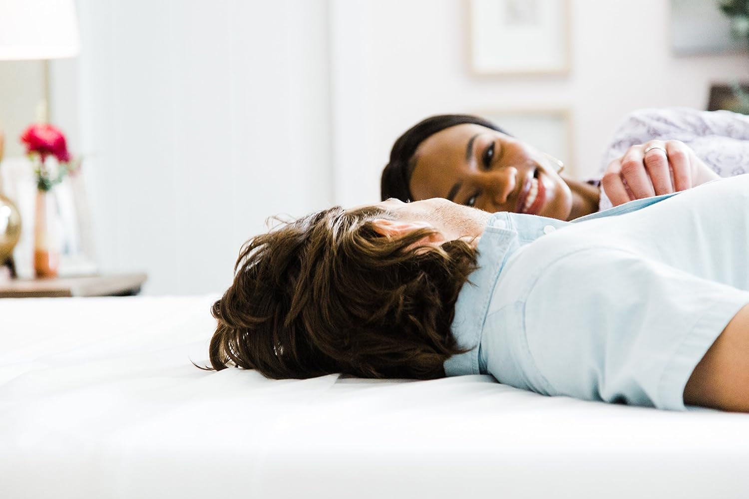Two people lying on a mattress, smiling and facing each other in a bright bedroom.