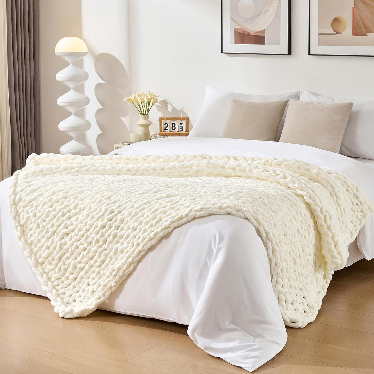 Cream chunky knit throw laid across a bed in a styled bedroom with pillows and lamp.