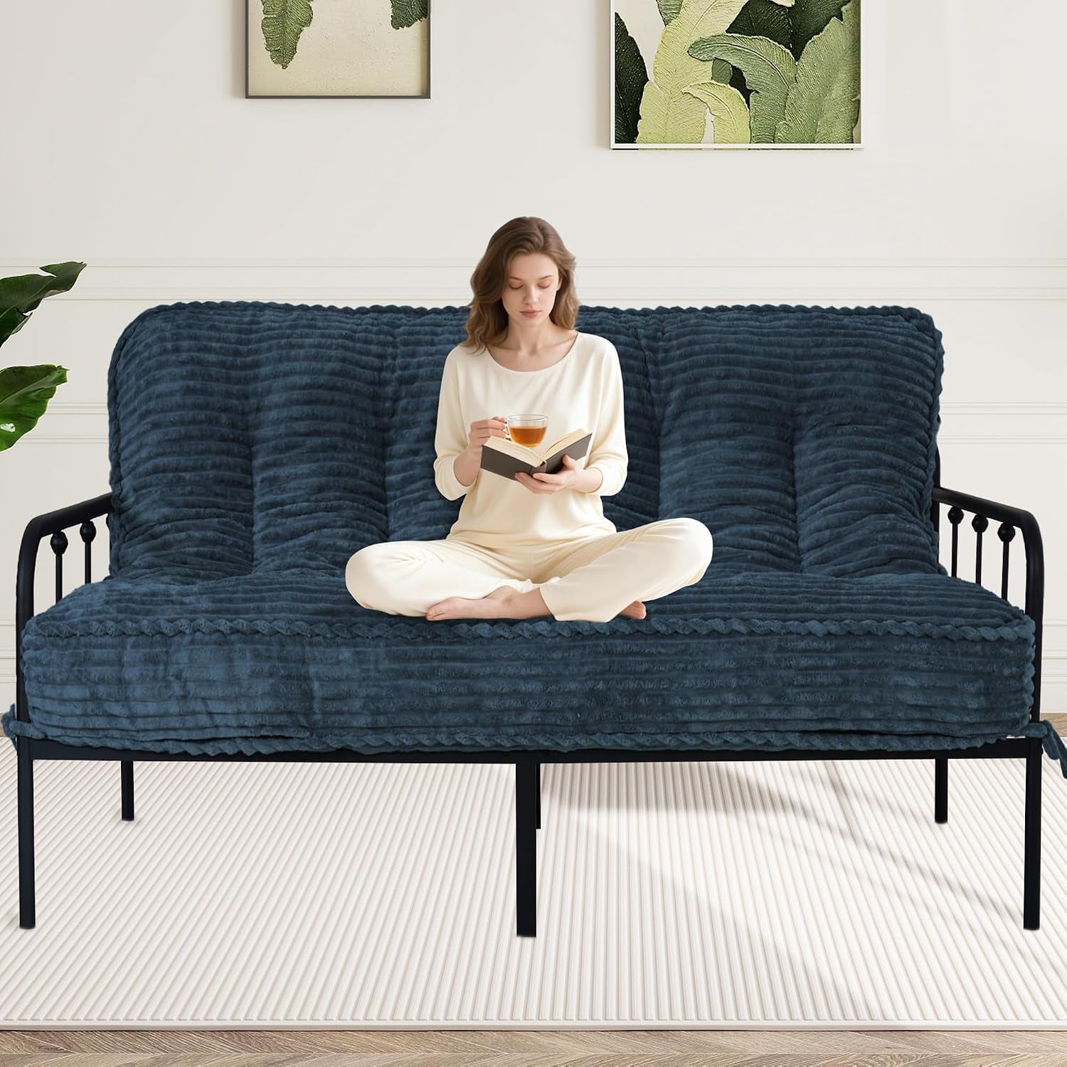 Front view of navy blue quilted futon mattress on black metal frame with woman sitting cross‑legged reading