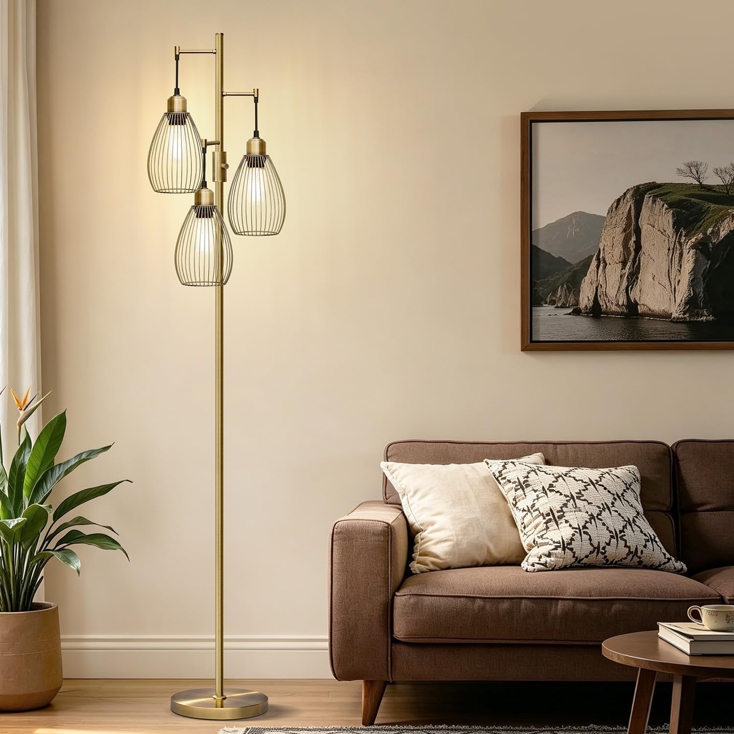 Gold floor lamp with three lit teardrop wire shades standing beside a brown sofa and framed landscape