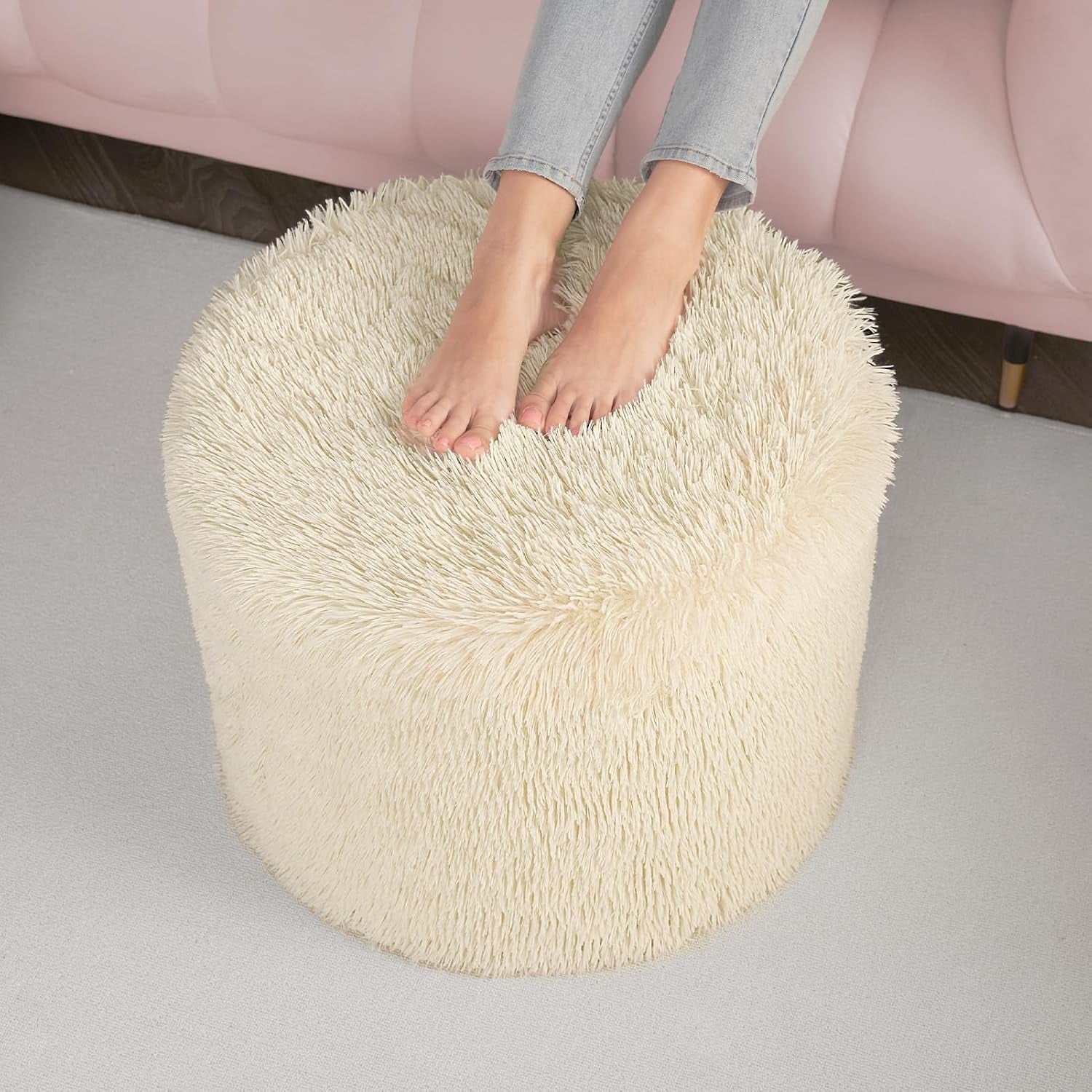 Beige round faux fur pouf shown from a high angle with feet resting on top next to a pink sofa