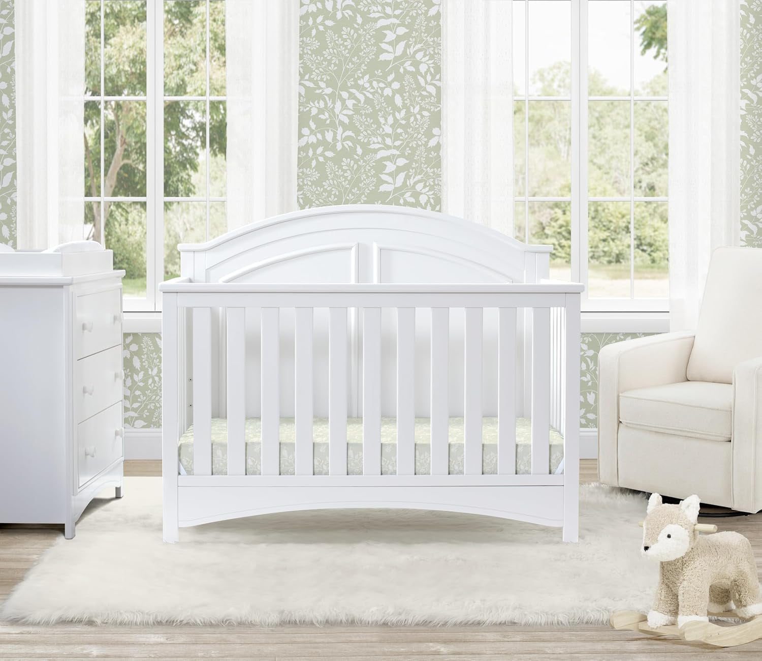 White crib staged in a nursery with dresser, armchair, rug and stuffed animal by a window.