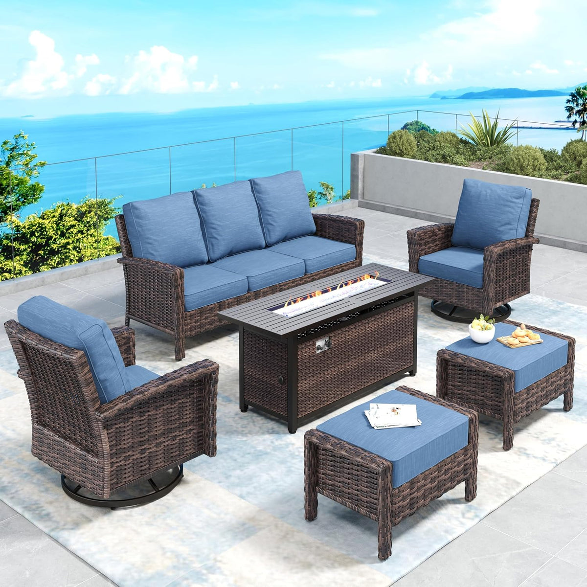 Patio lounge set with blue cushions, fire pit table center and two wicker ottomans on a pale rug with ocean backdrop.