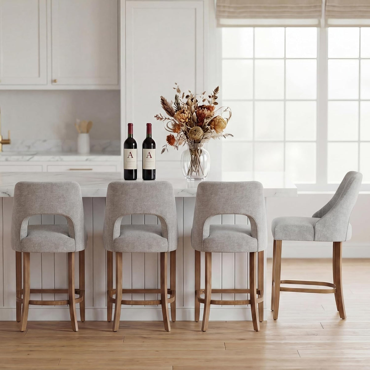 Four light gray upholstered counter stools with wood legs arranged at a kitchen island under a large window.