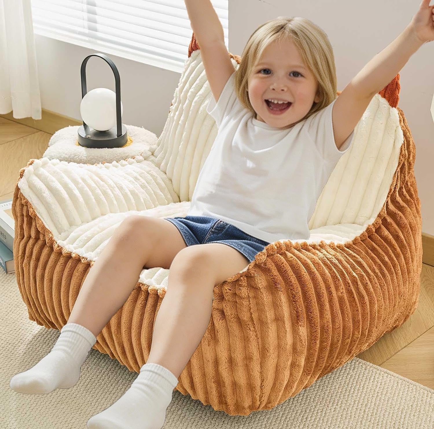 Child with arms raised sitting on a two-tone brown corded bean bag with cream ribbed interior and small lamp nearby.