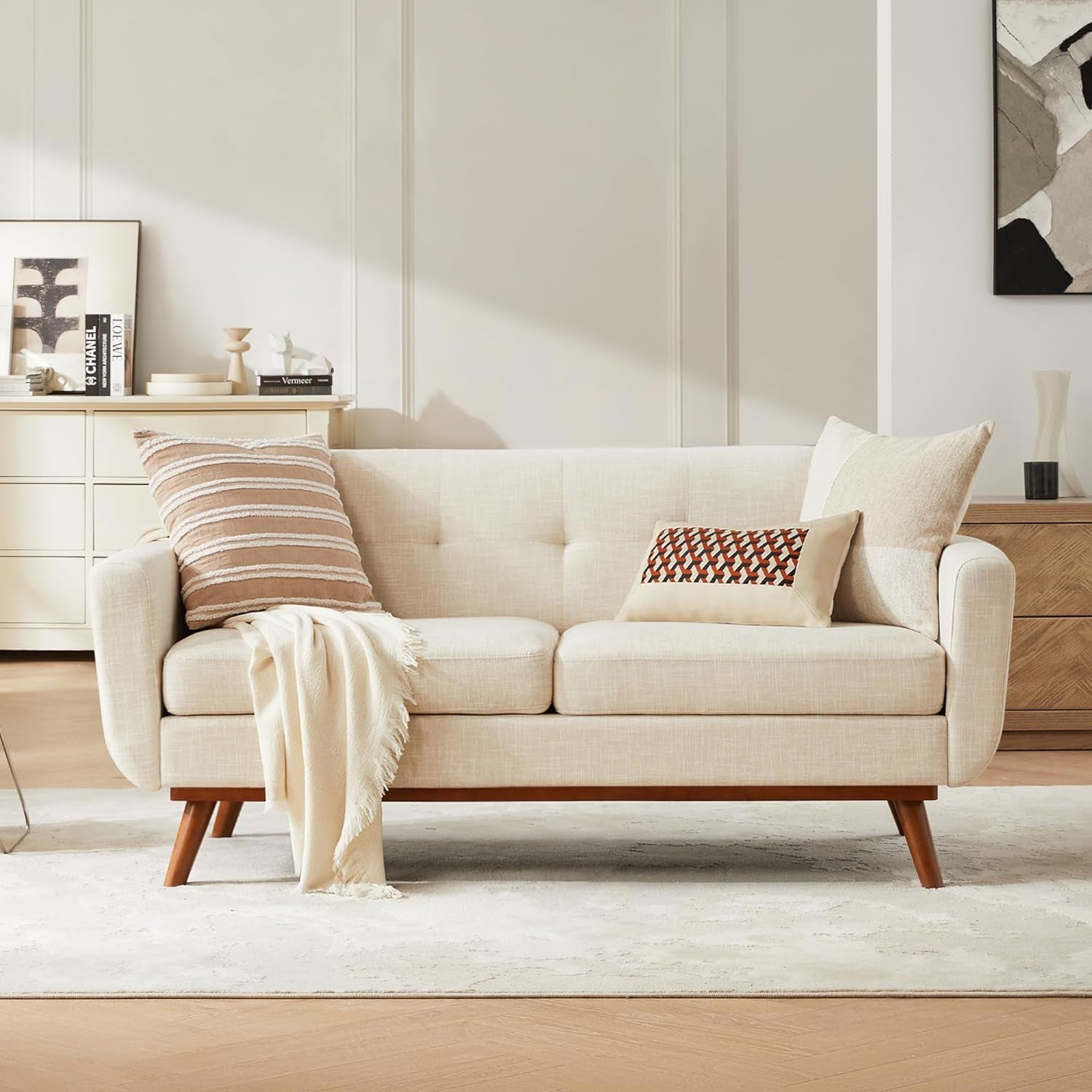 Beige two-seat sofa with button-tufted back, wooden tapered legs, throw pillows and a draped blanket in a living room.