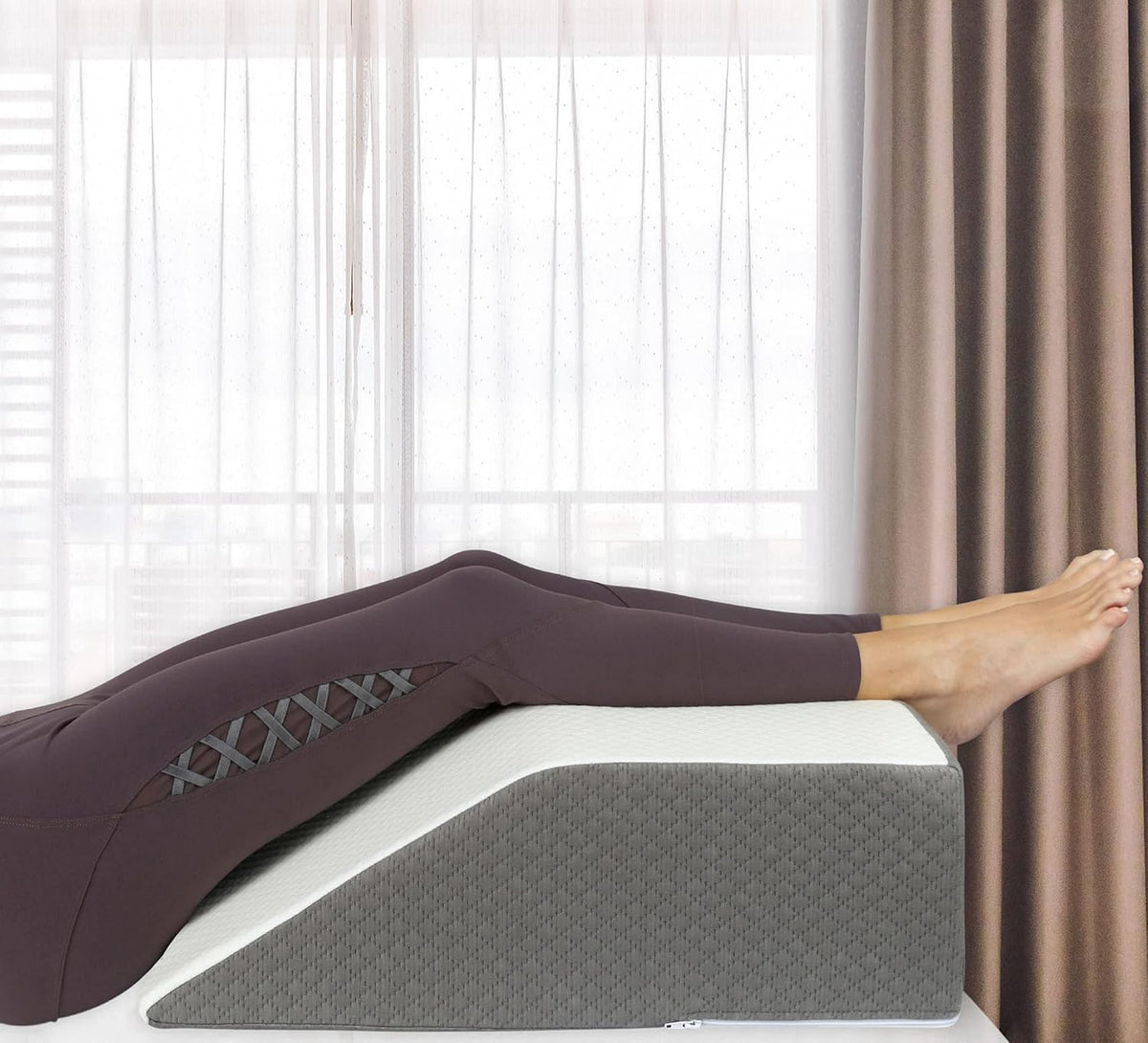 Close-up of legs elevated on the wedge showing the pillow's slope and quilted gray side against sheer curtains.