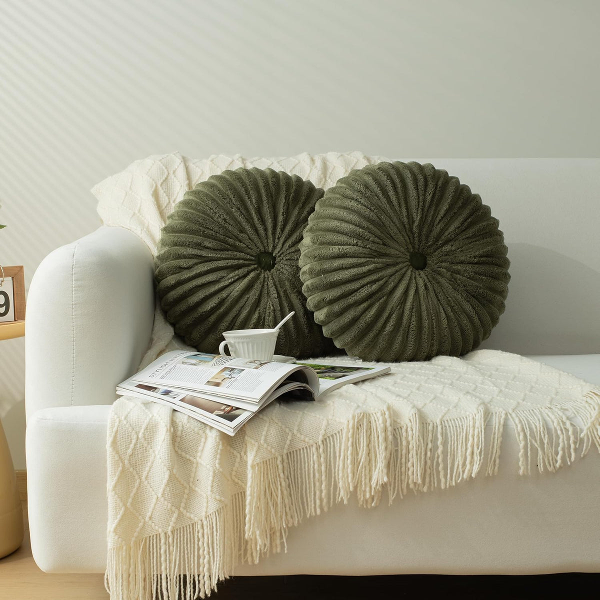 Two round olive-green ribbed pillows on a sofa draped with a cream throw and an open magazine.