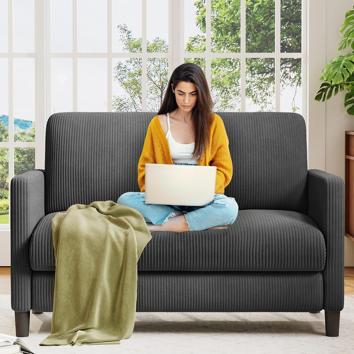Front view of a dark gray ribbed loveseat with a woman using a laptop and a green throw draped over the seat.