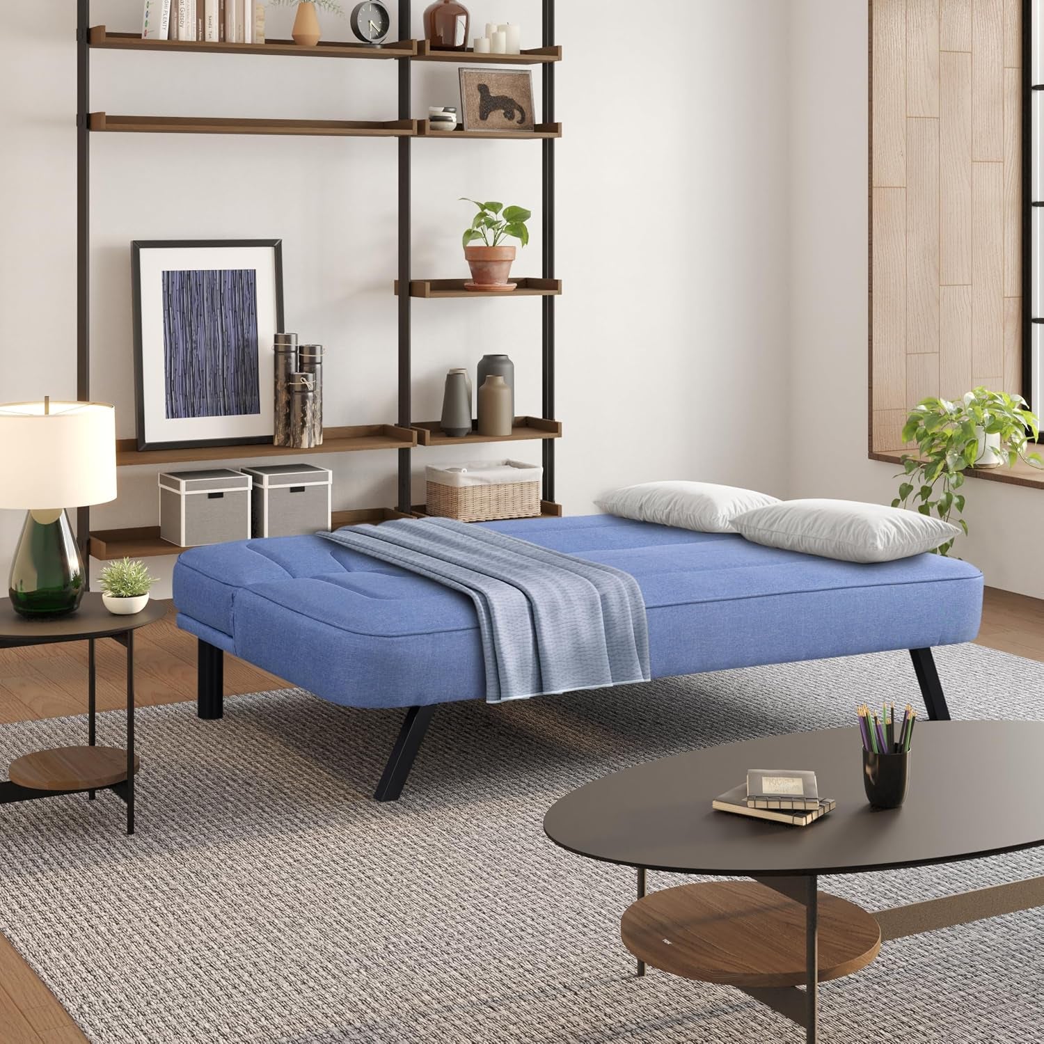 Blue upholstered futon shown converted to a flat bed with a blanket and pillows in a living room