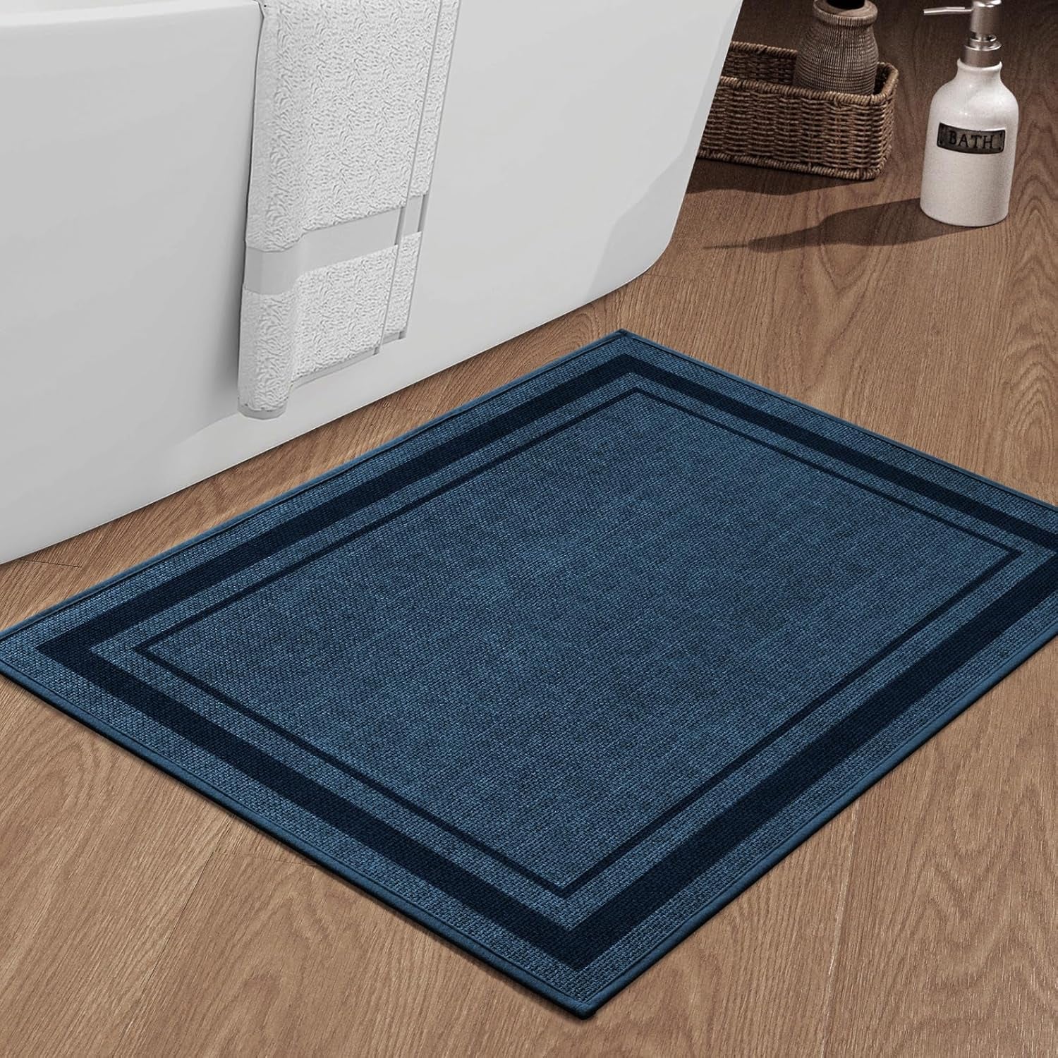 Square dark blue bath mat on wood floor in front of a bathtub with towel and bath accessories nearby