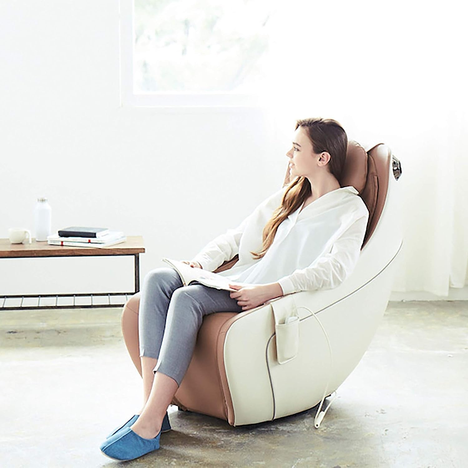 Young woman reclining in a beige-and-white massage chair reading a book in a sunlit room.