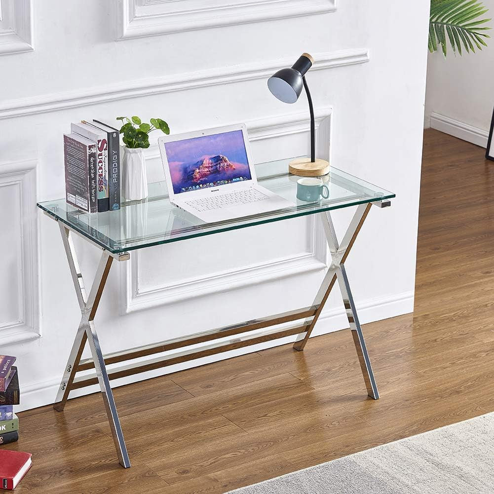 Frontal view of the glass desk against a white paneled wall with books on the left, laptop center, and lamp on the right.
