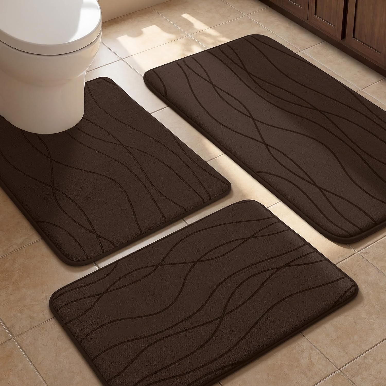 Three dark brown memory-foam mats with flowing embossed lines arranged on a tiled floor near a toilet and cabinet.