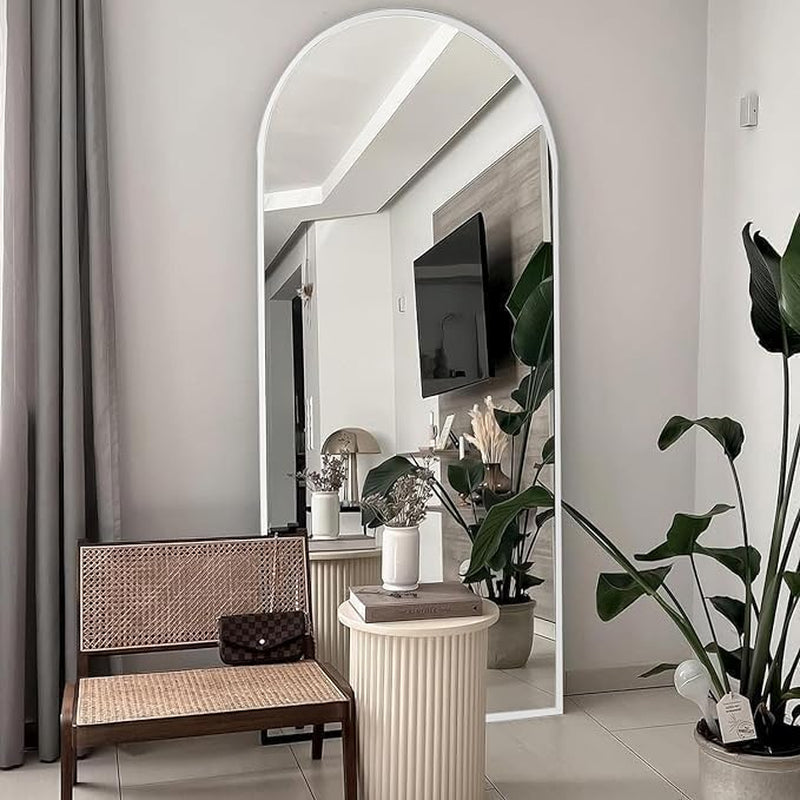 Arched full-length mirror with slim white frame leaning against a wall, reflecting a chair and plants.