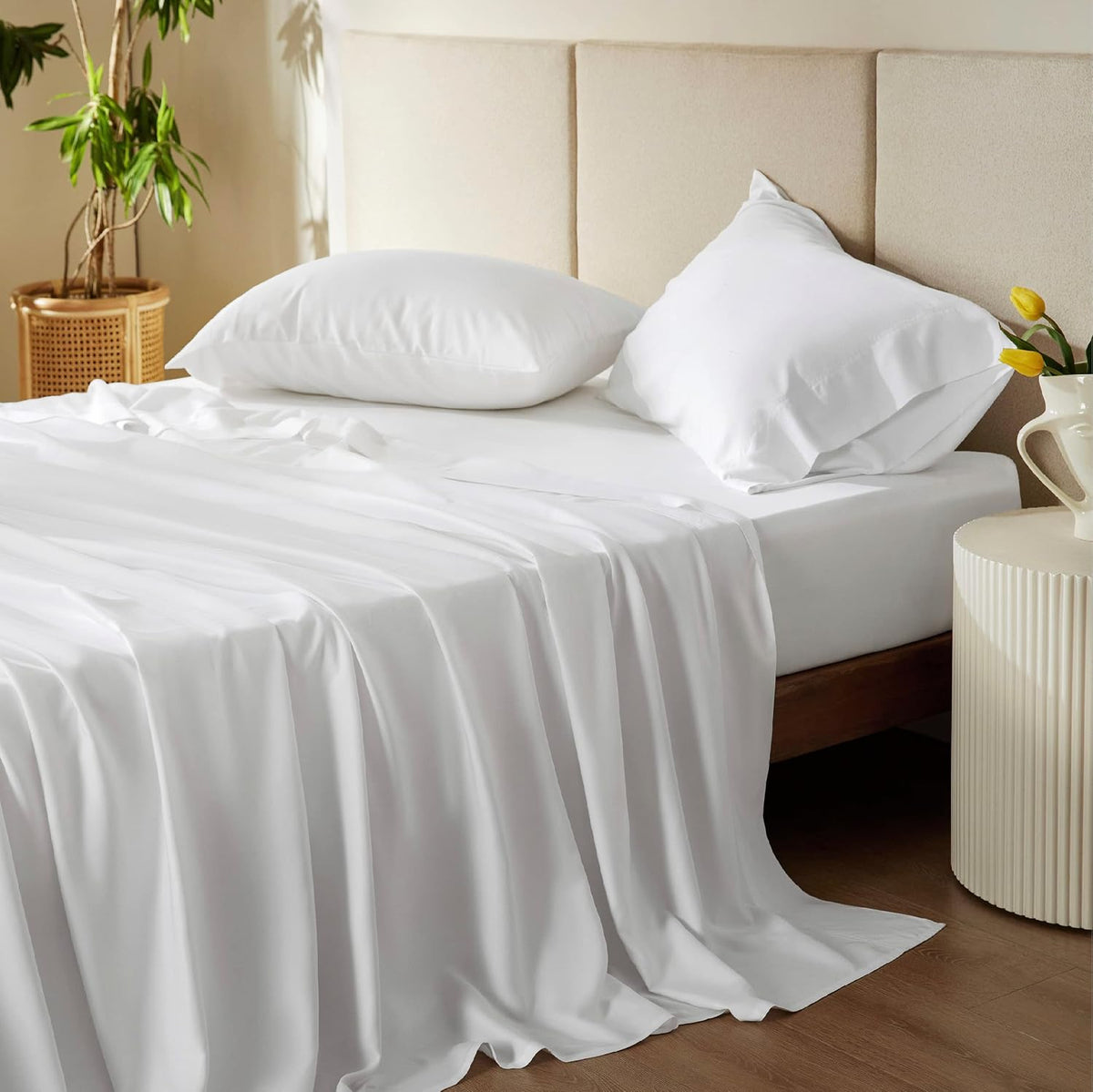 Front view of a made bed with white sheets, two pillows, beige upholstered headboard and a round side table with flowers.