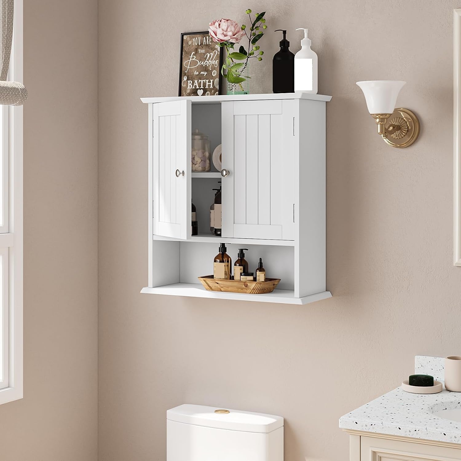 White over-toilet cabinet mounted on a beige wall with decorative items on top and toiletries on the open shelf.