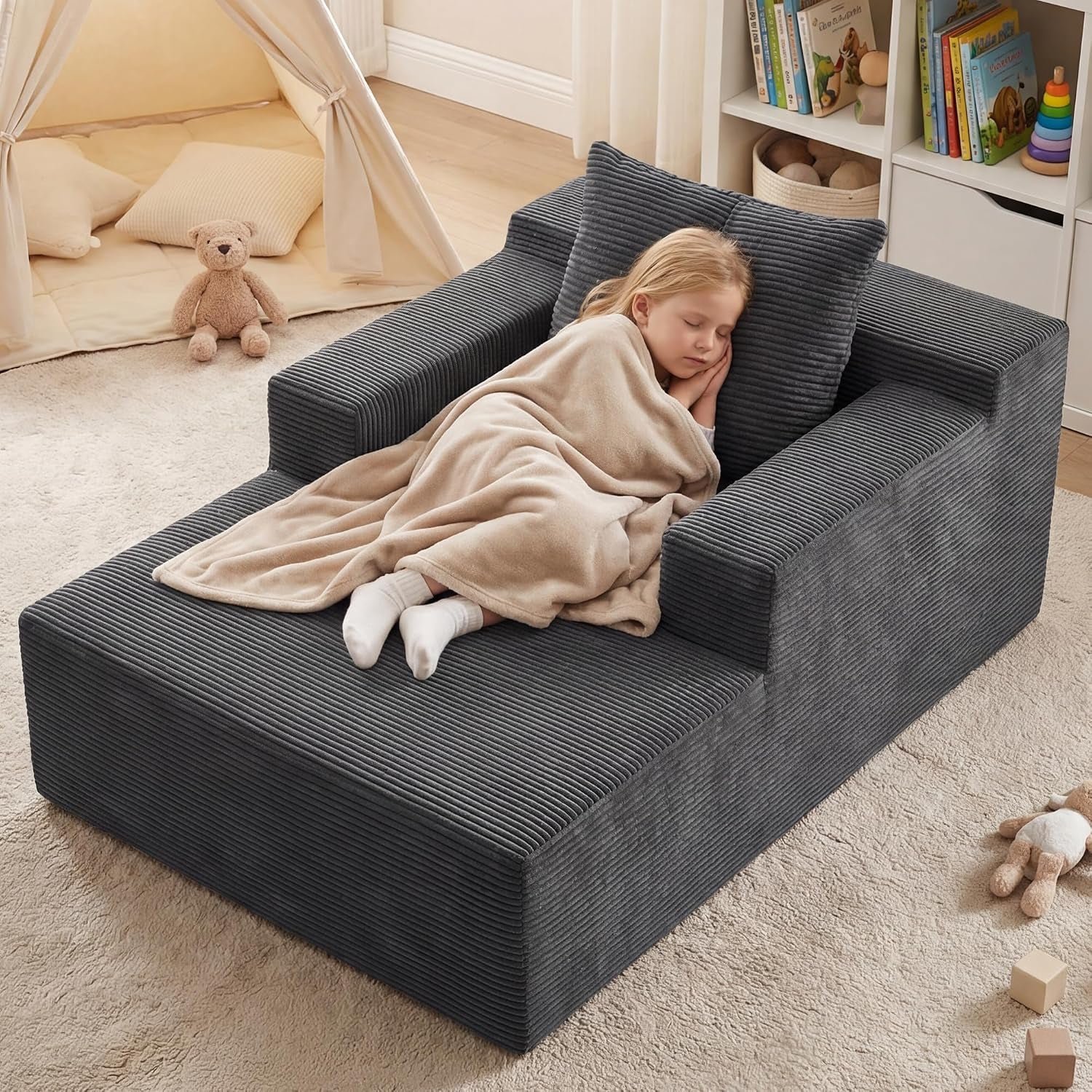 Child wrapped in a beige blanket sleeping on a dark gray corduroy chaise lounge in a playroom with a toy tent and shelving.
