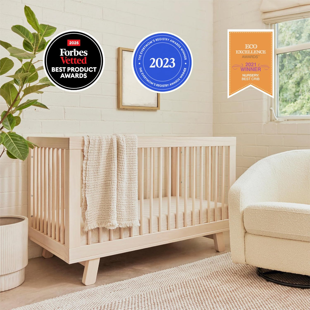 Washed natural wooden crib in a styled nursery with a cream blanket draped over the rail and an armchair nearby.