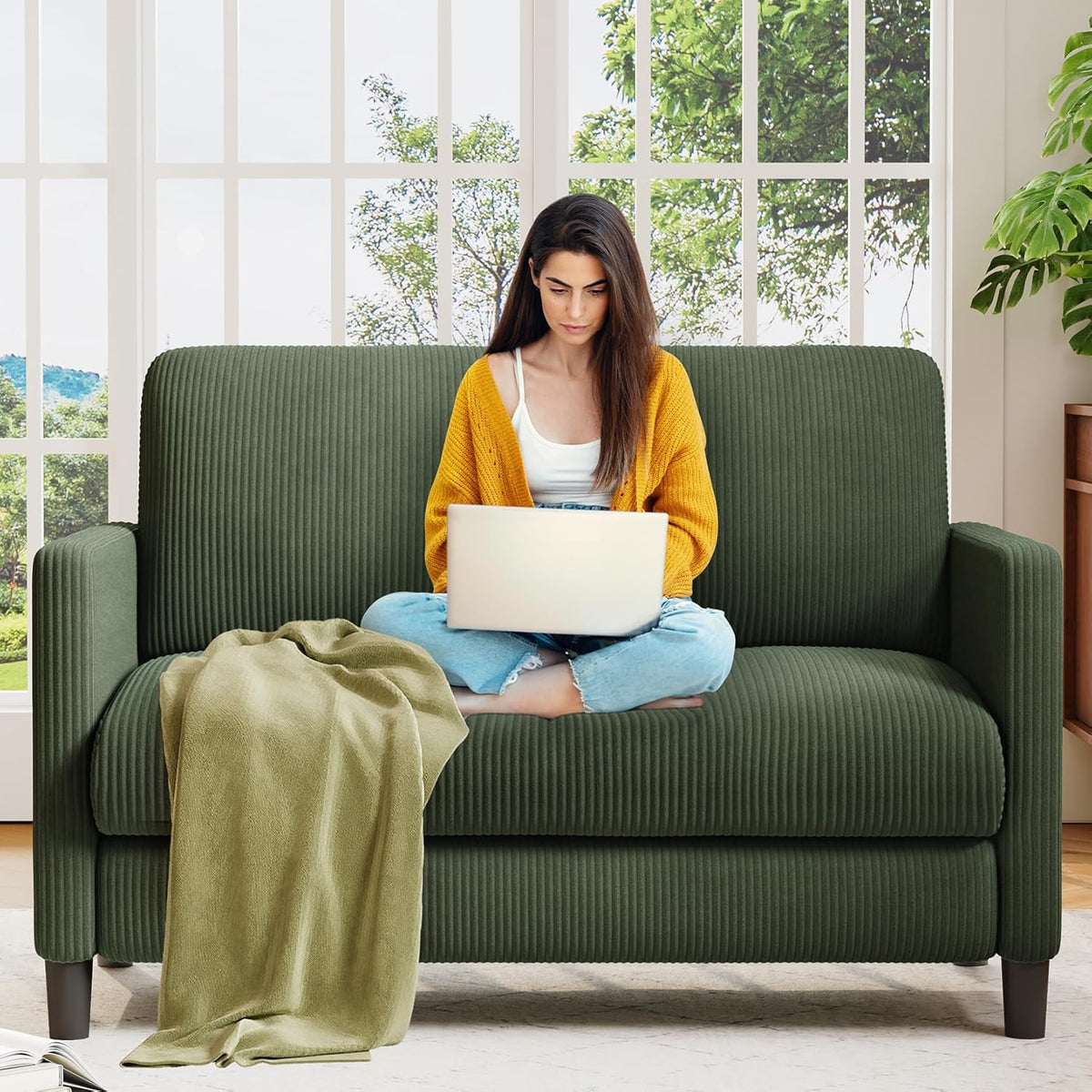 Front view of a small dark green ribbed loveseat with a woman sitting cross‑legged using a laptop and a green throw on the
