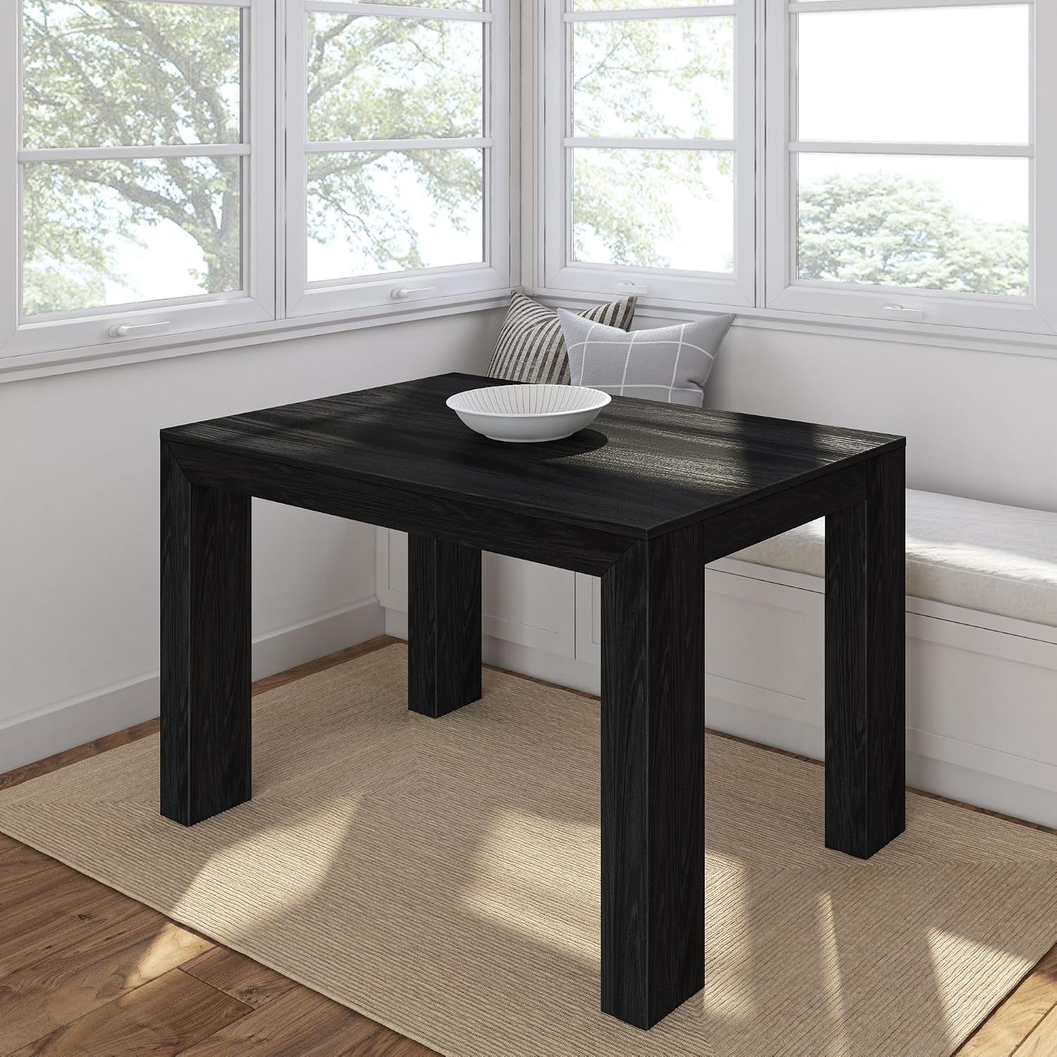 Square black wood dining table in a bright windowed breakfast nook with a bowl on top.