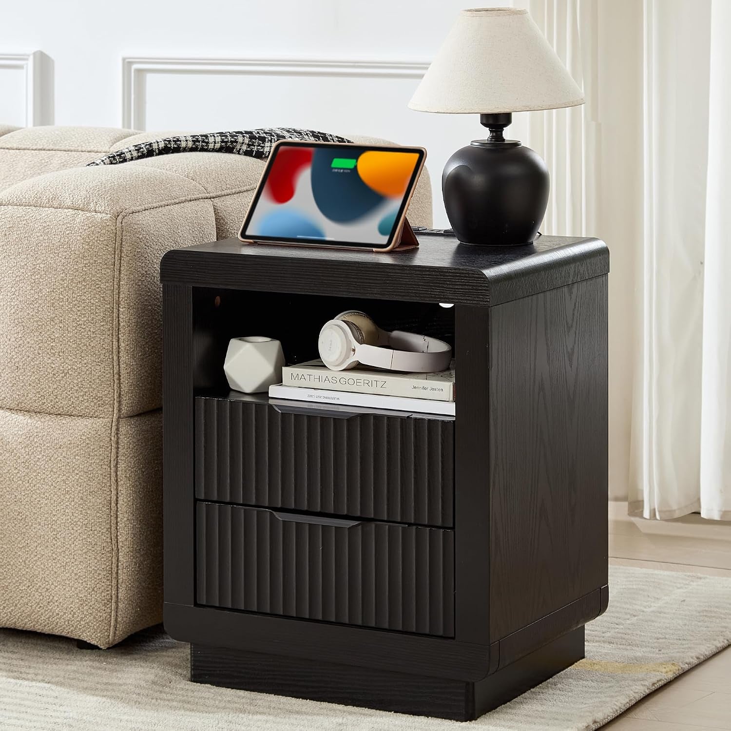 Single dark wood nightstand next to a beige sofa holding a lamp, tablet on top and headphones and books in the open shelf.