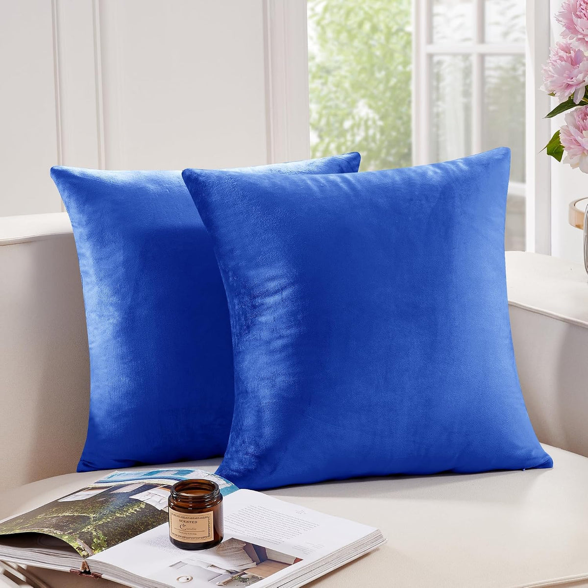 Two square bright blue throw pillow covers on a beige sofa next to a candle and open book