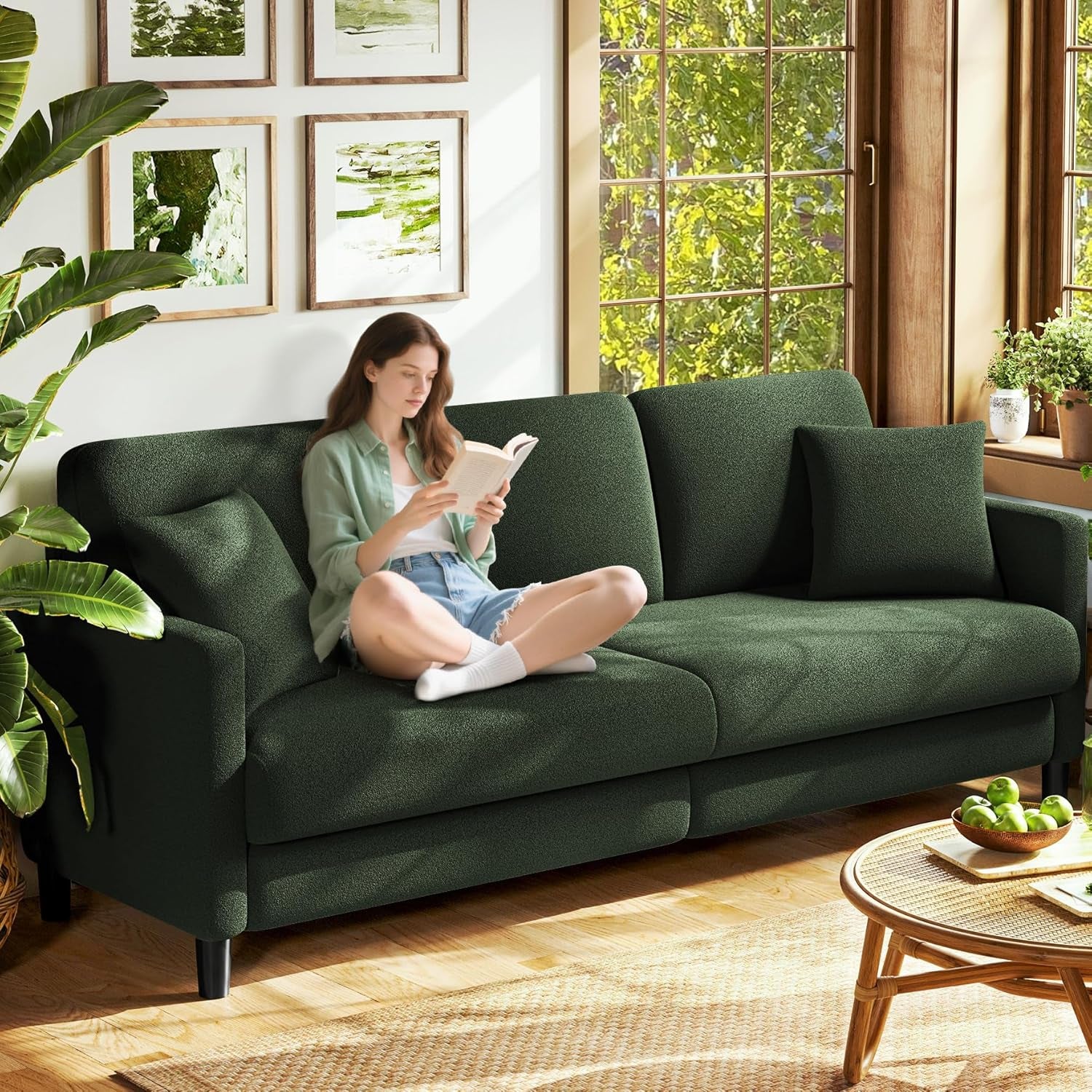 Dark green two-seat sofa by a large window with a woman sitting cross-legged reading a book.