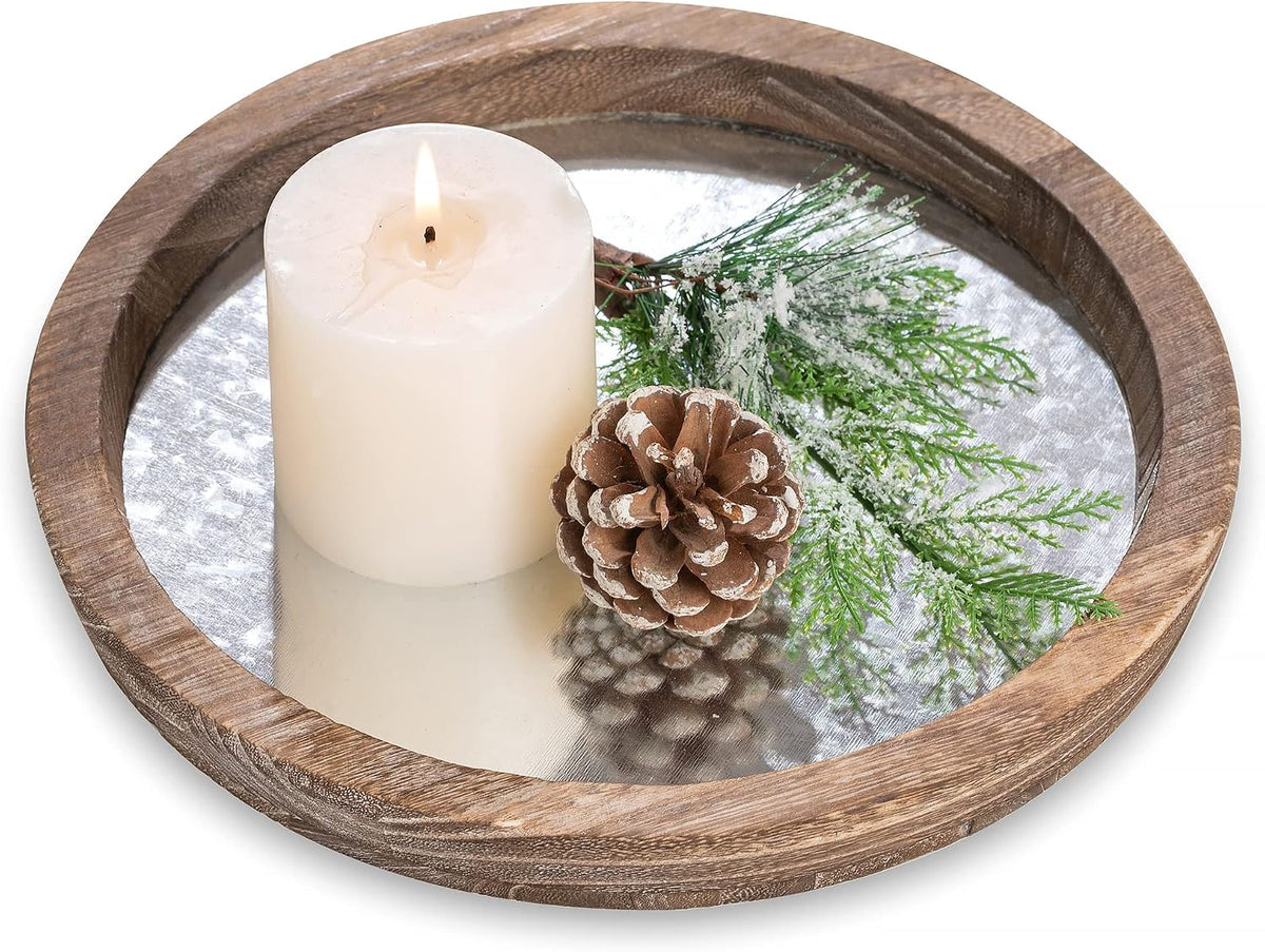 Round wooden tray with mirrored metal center holding a lit pillar candle, pinecone and frosted greenery