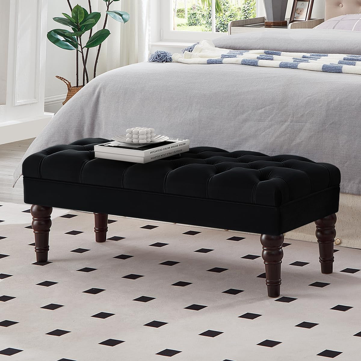 Black button-tufted bench with turned dark legs beside a bed, a small decorative tray and books on the seat.