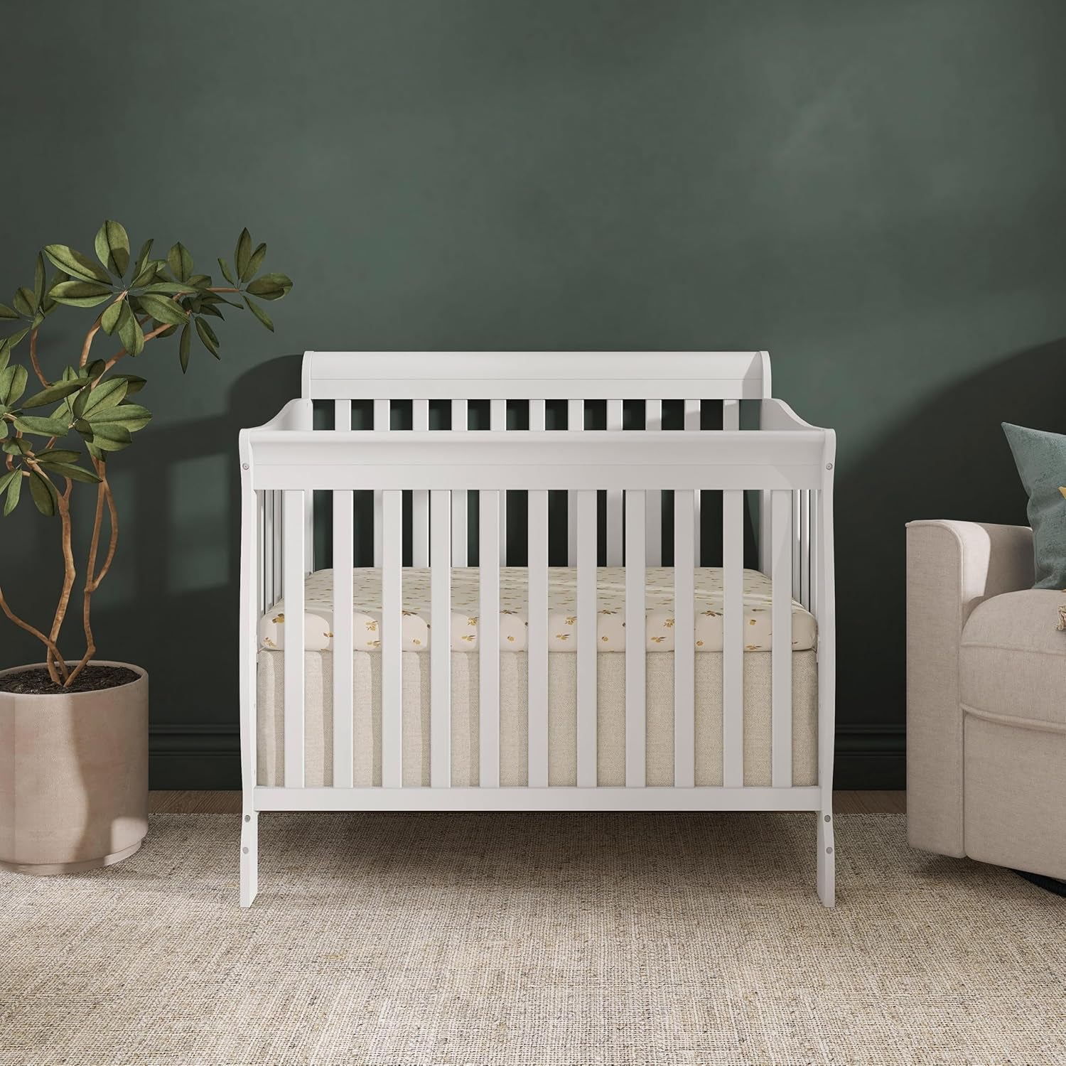White mini crib placed against a dark green wall between a potted plant and a beige armchair on a woven rug.