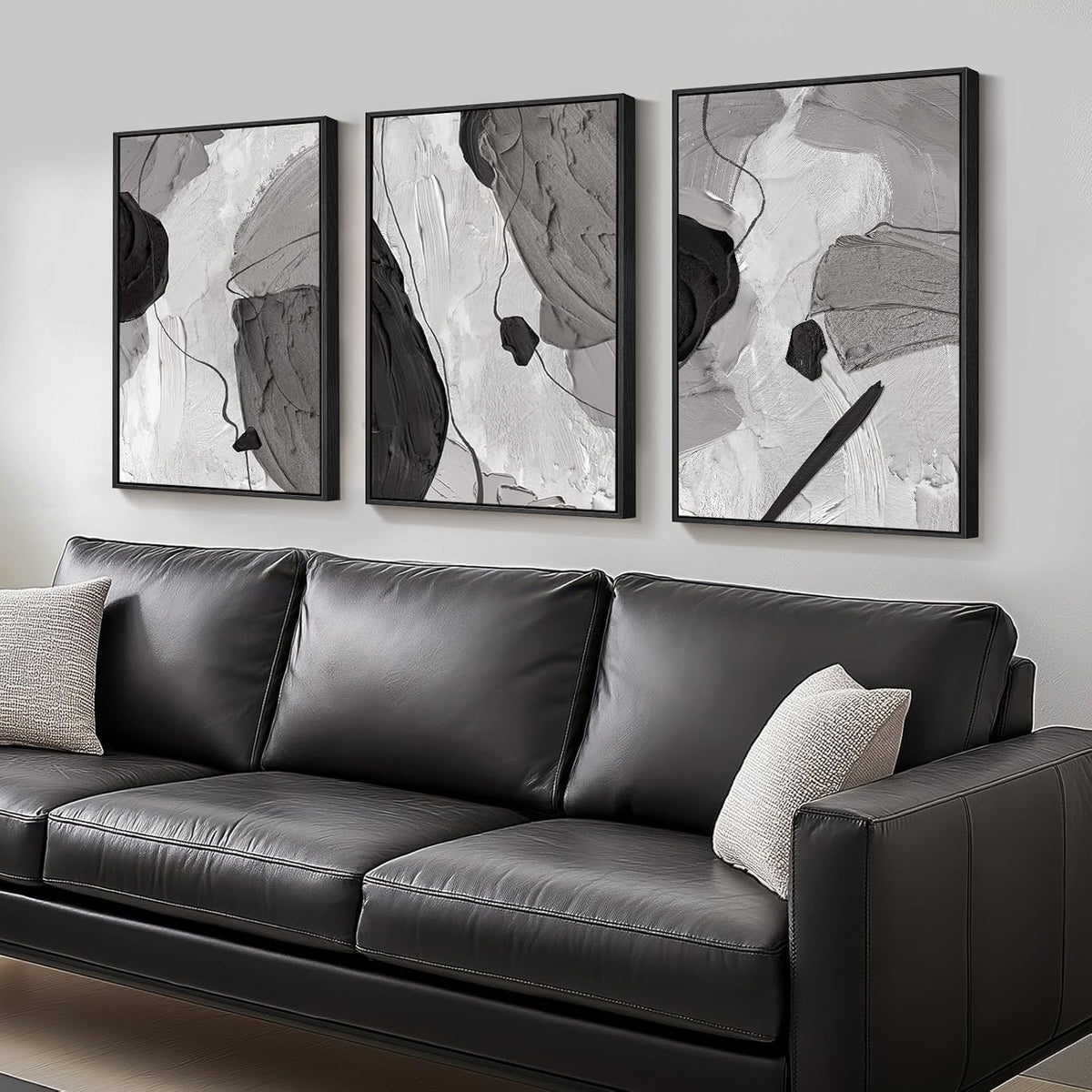 Angled side view of three tall black-framed abstract canvases mounted above a smooth dark sofa with a textured lumbar pillow.