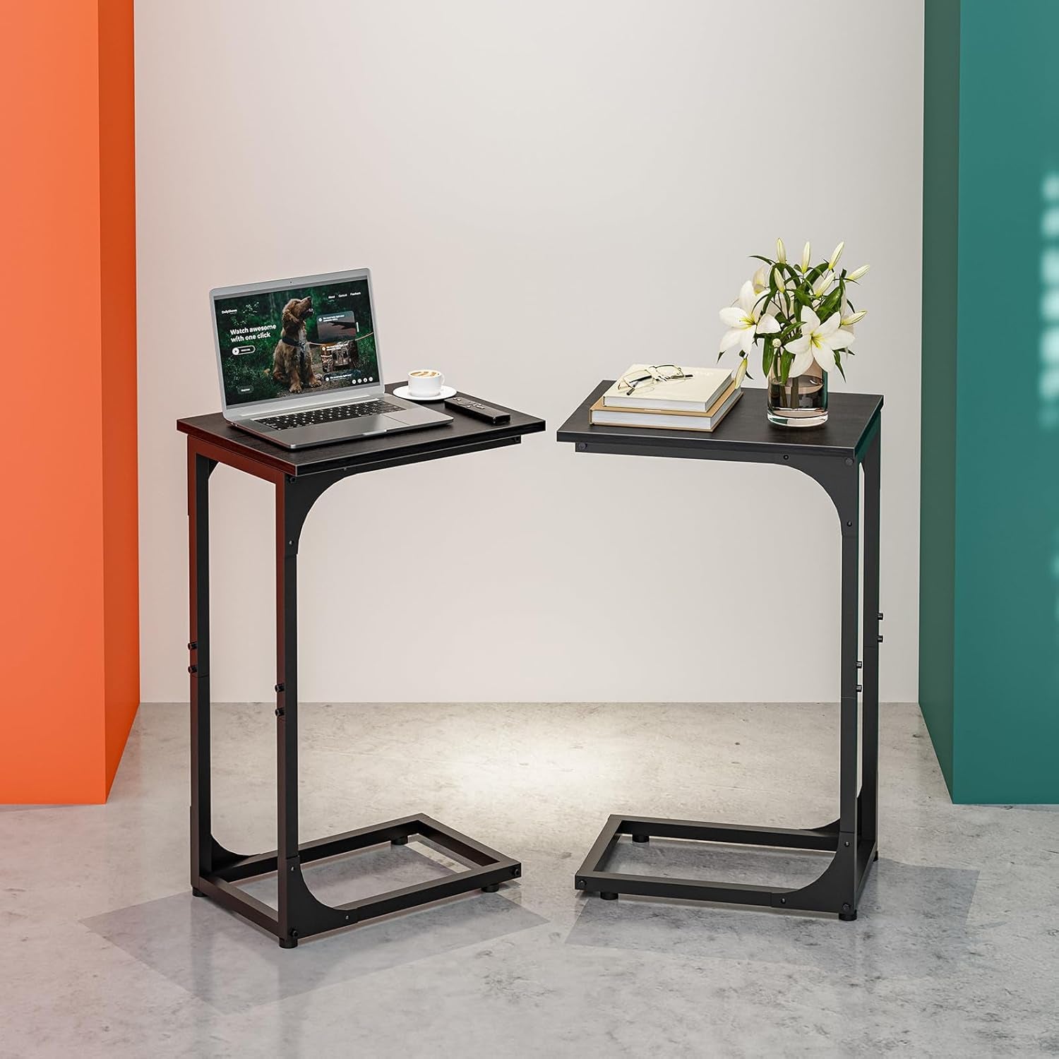 Two C-shaped end tables side by side with dark tops and black frames, one holding a laptop and the other books and flowers.