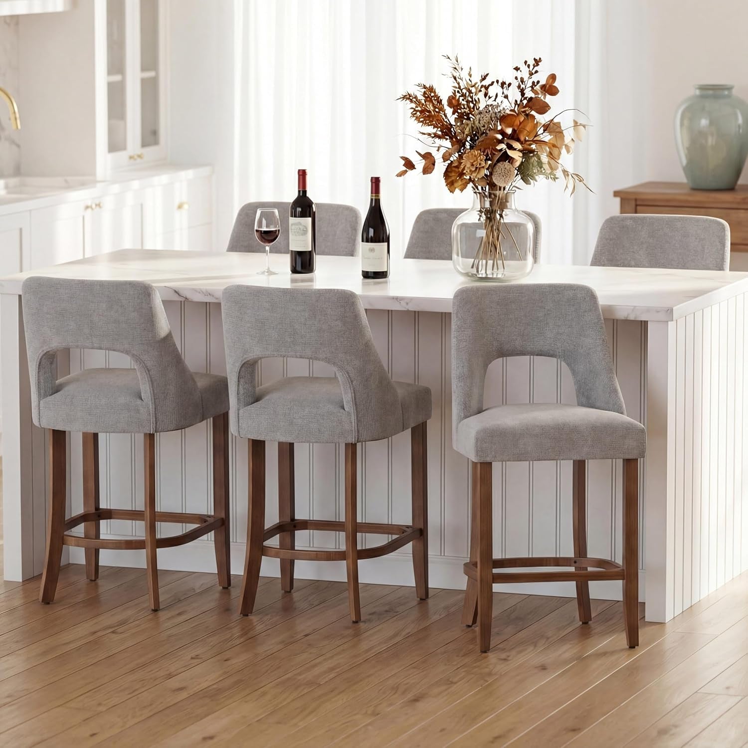 Four light-gray upholstered counter stools with wooden legs arranged along a kitchen island with vase and wine