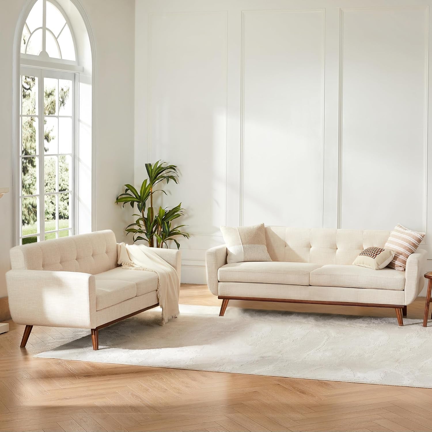 Beige loveseat and matching three-seat sofa styled in a bright room with tall window, rug, and plant